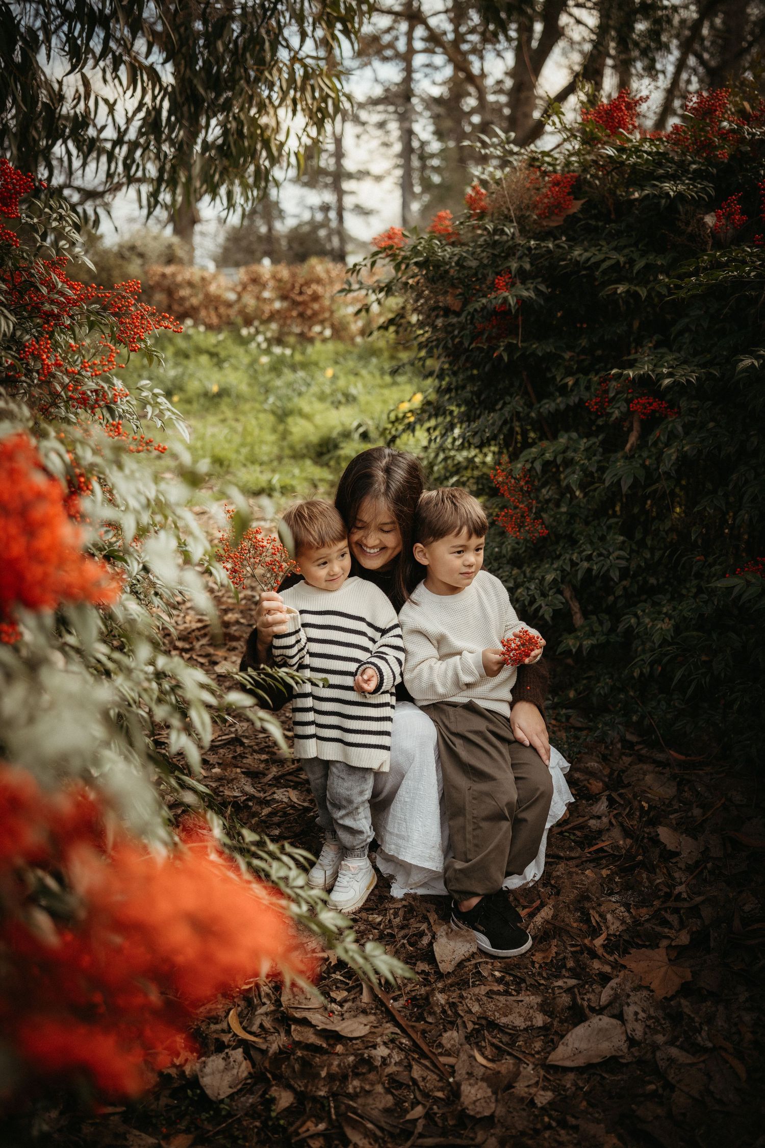 Mother embracing two young children among vibrant red berry bushes in a lush garden setting.
