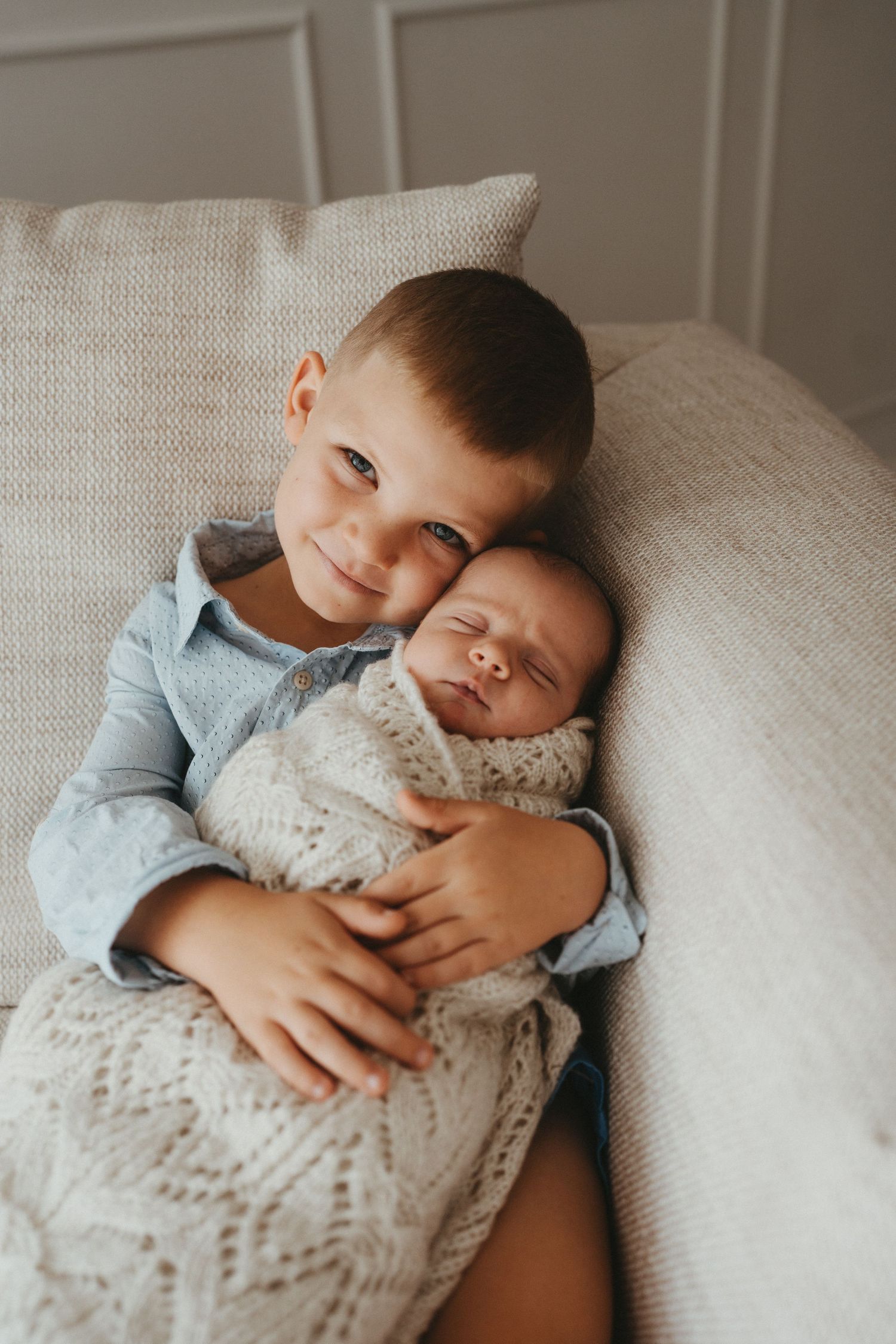 Young boy gently holding sleeping newborn baby sibling wrapped in knit blanket on couch.