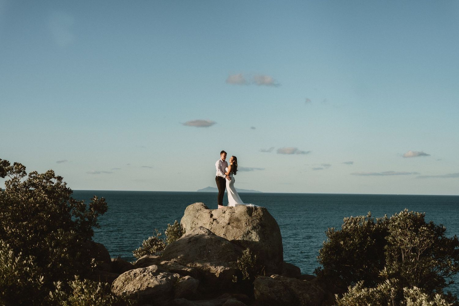 Couple in wedding attire standing on coastal rocks surrounded by lush greenery with ocean and blue sky backdrop.