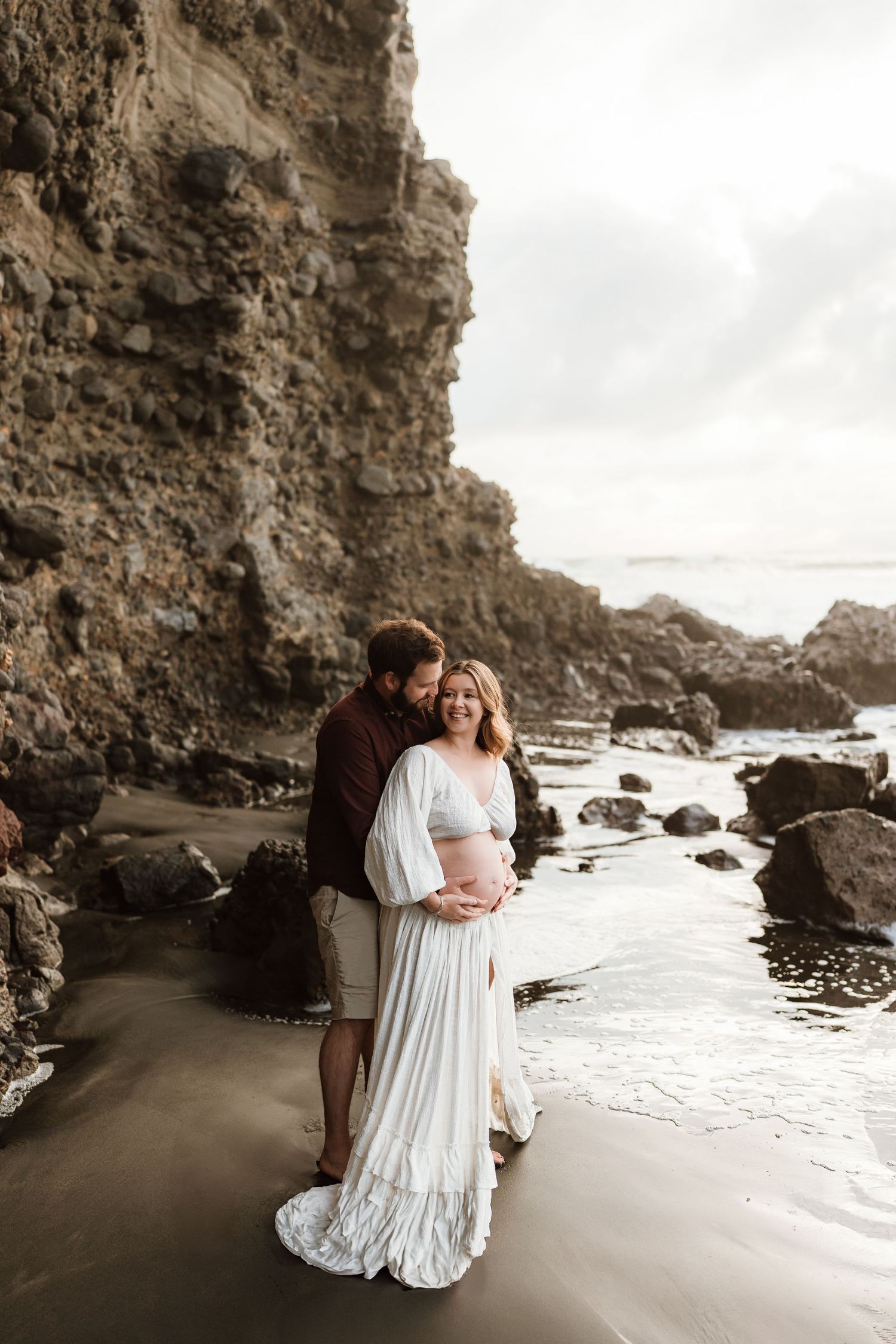 Couple poses on rocky beach, pregnant woman in white dress embraced by partner near coastal cliffs.