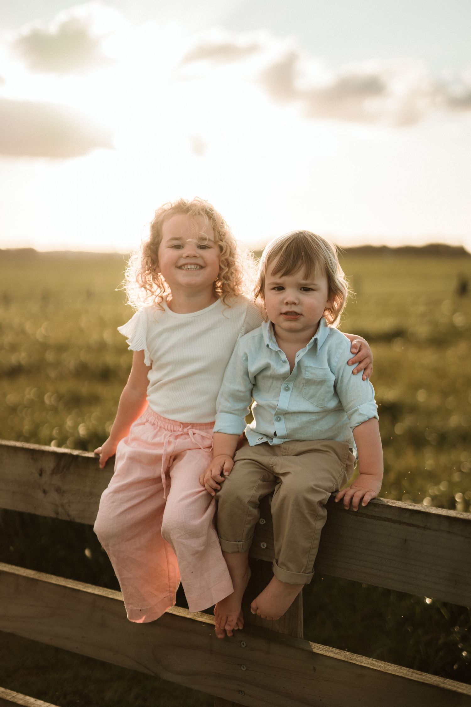 Two young siblings sitting on a wooden fence at golden hour in a green field, smiling for a portrait. Katie Charlotte Photography