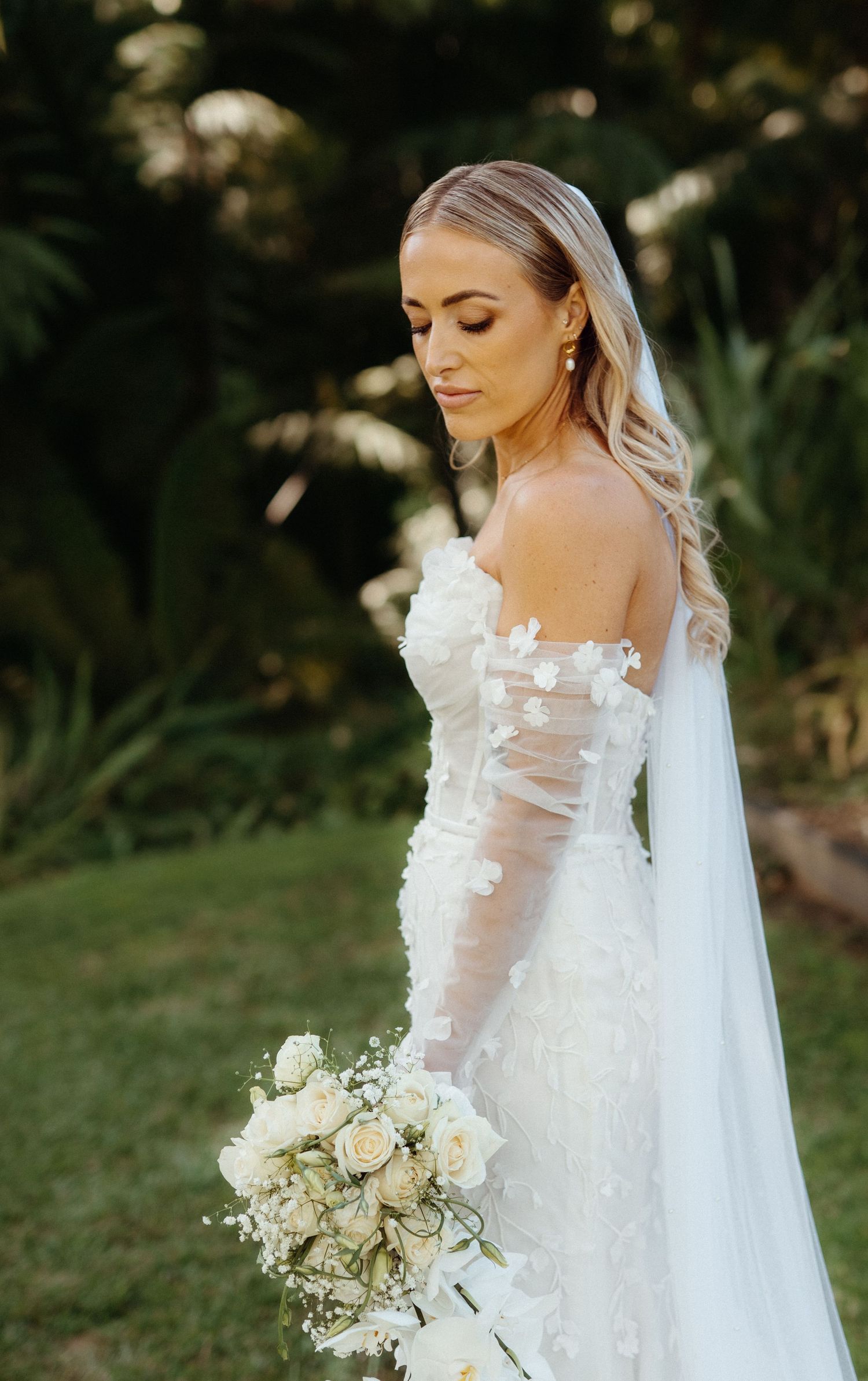 Elegant blonde bride in off-shoulder lace gown with veil holding a white rose bouquet outdoors.