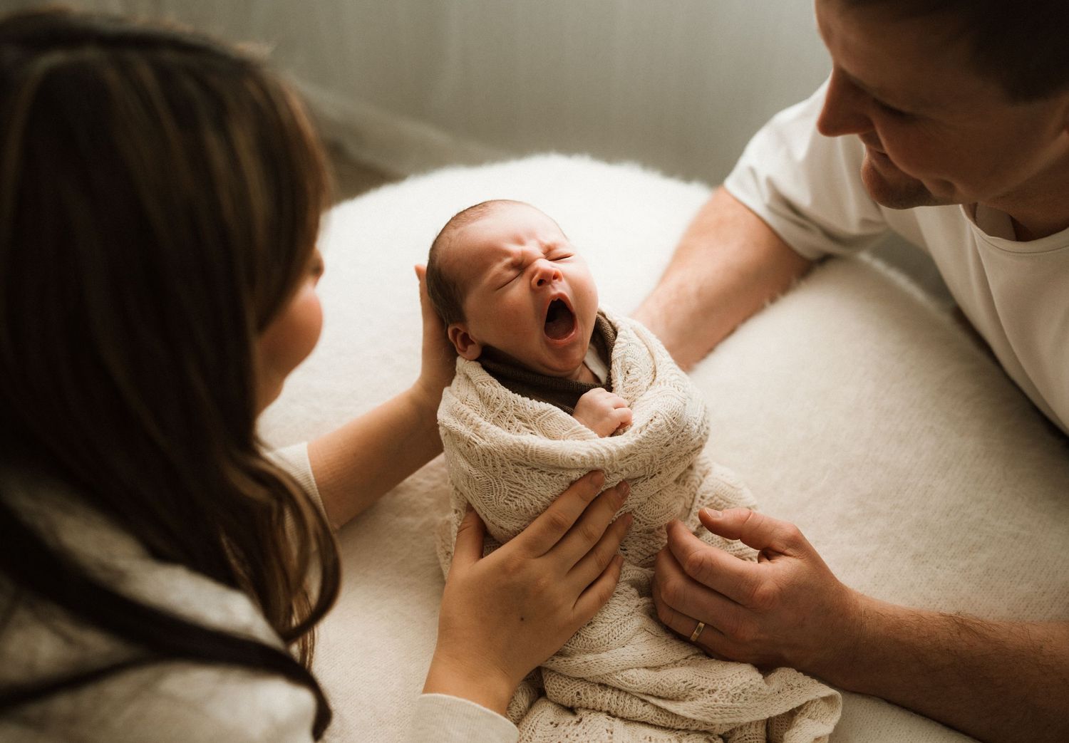 Newborn baby yawning while swaddled in white blanket, held by two parents smiling lovingly.