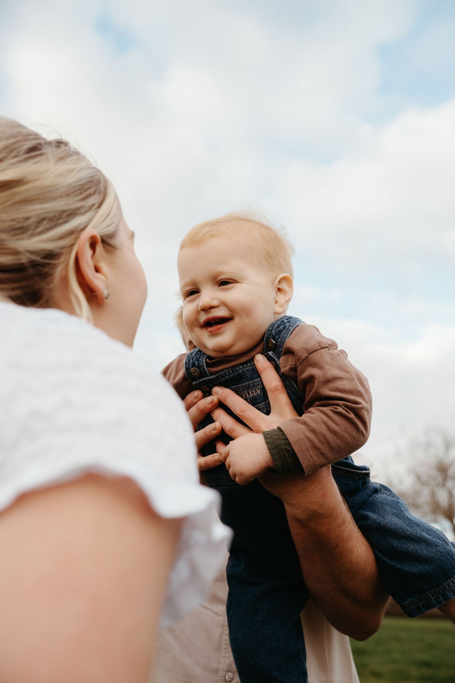 A joyful baby smiles while being held up by a parent outdoors under a cloudy sky.
