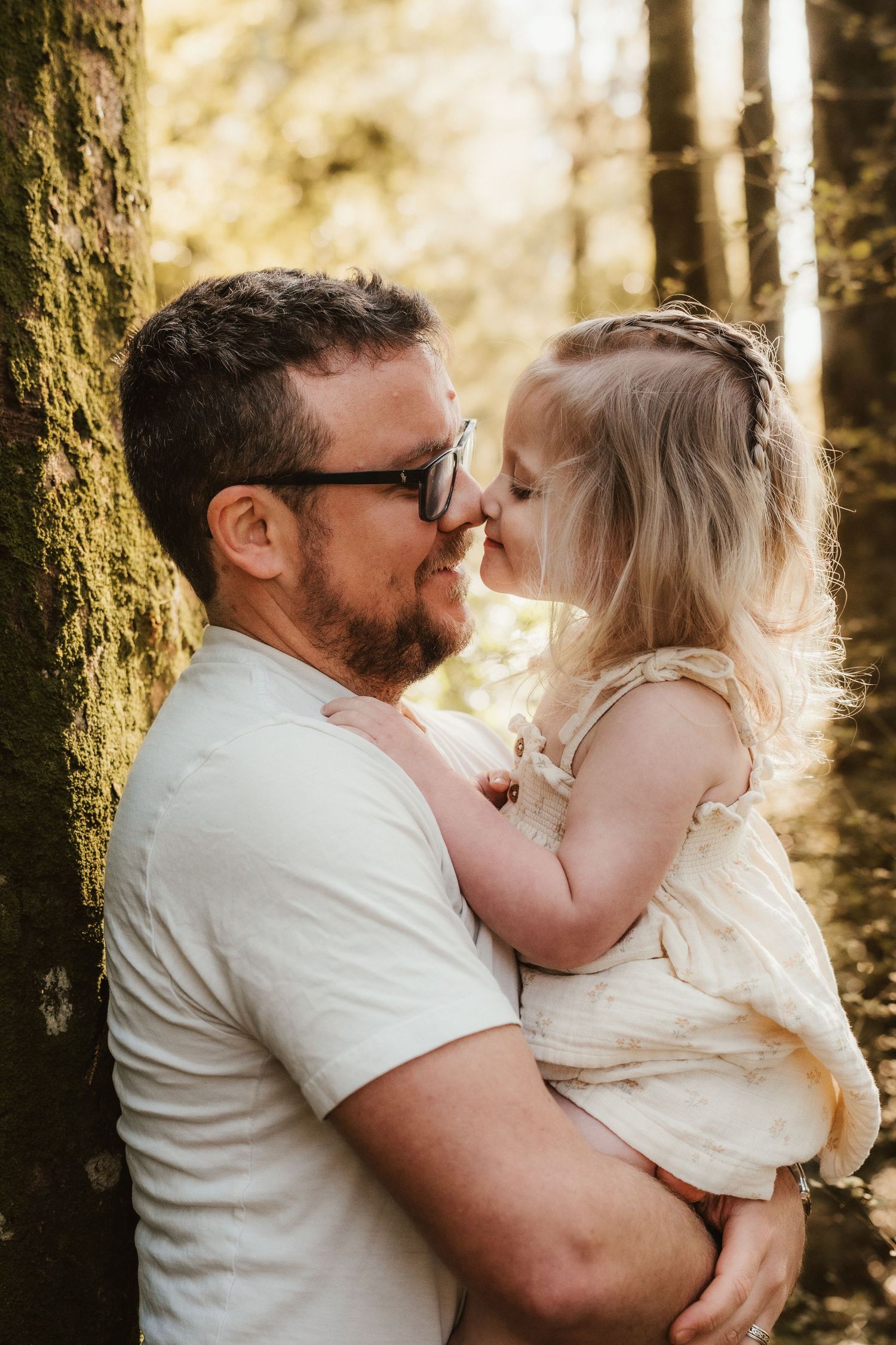 Father holding young daughter nose-to-nose in sunlit forest, sharing tender moment beside moss-covered tree.