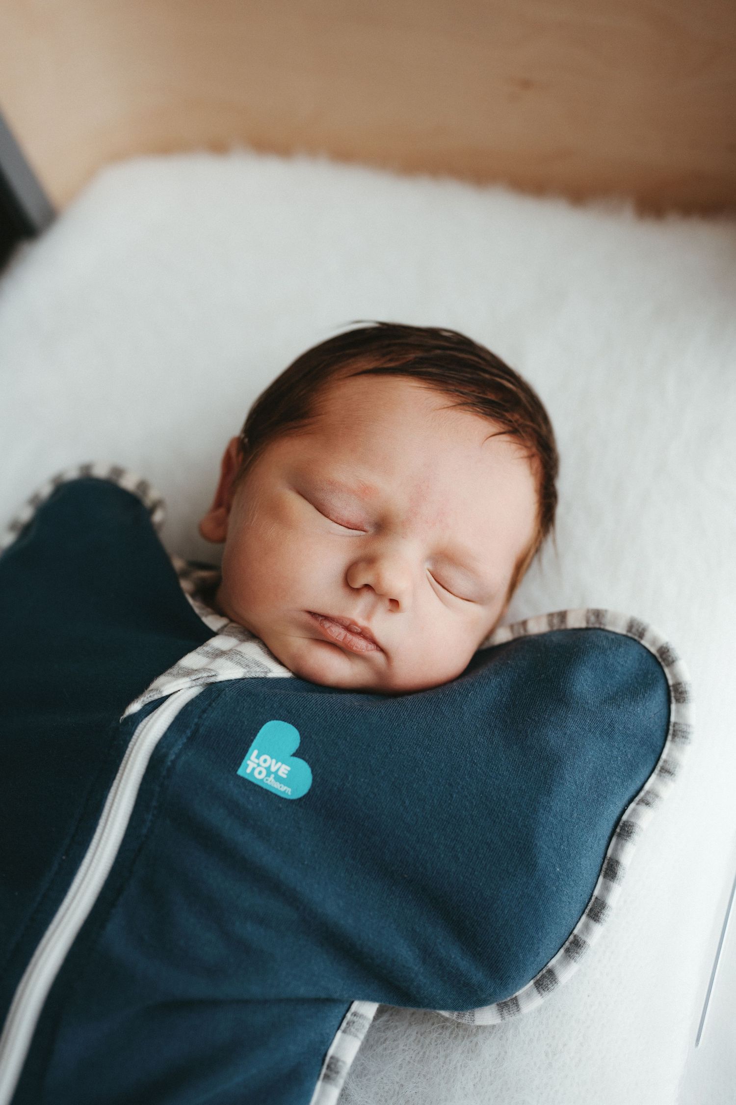 Newborn baby sleeping peacefully in a navy Love to Dream swaddle wrap inside a white fleece-lined bassinet.