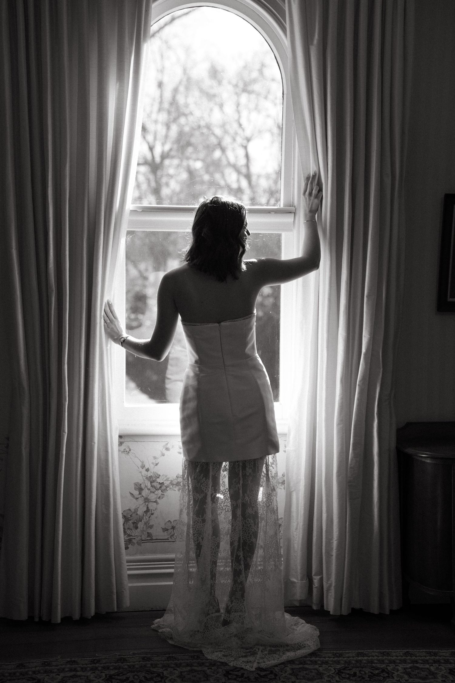 Black and white photo of a bride in a lace wedding dress standing at an arched window with curtains.
