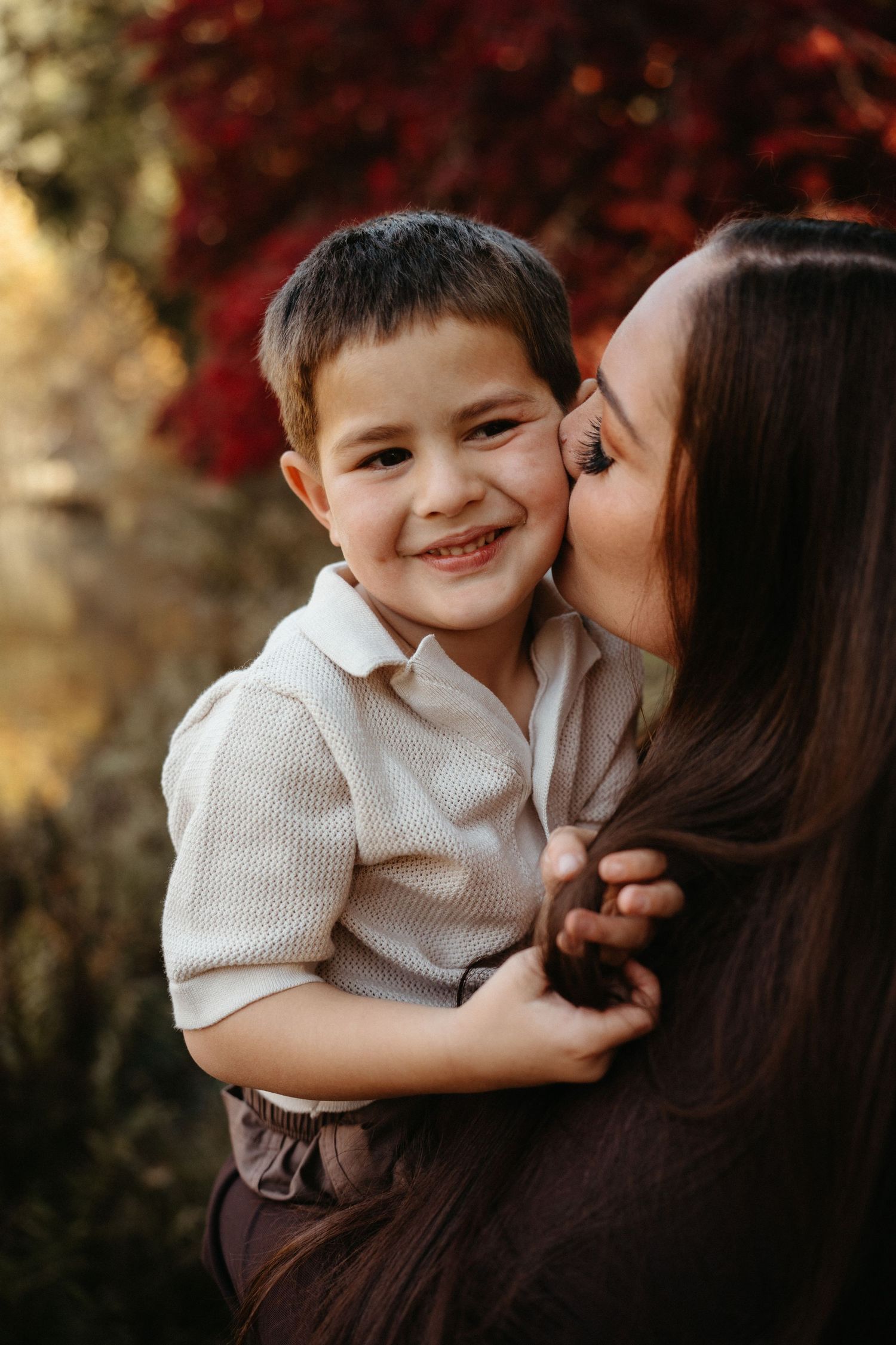 A mother kisses her smiling young son on the cheek while holding him outdoors among red autumn foliage.