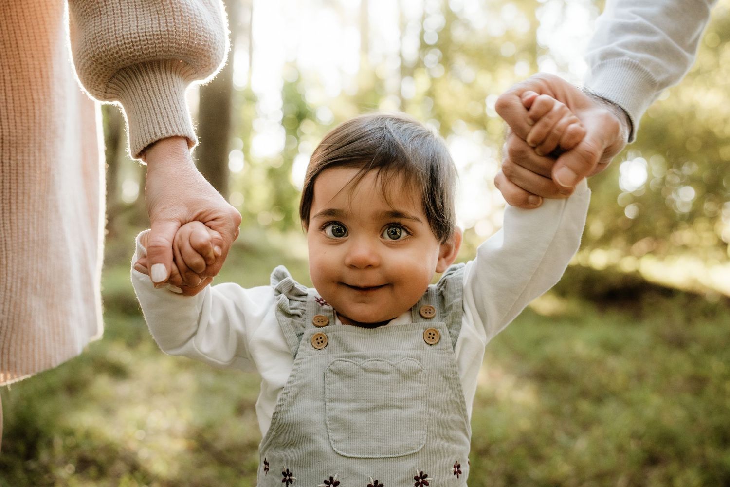 Adorable toddler girl in overalls smiling while being held up by both parents hands outdoors.