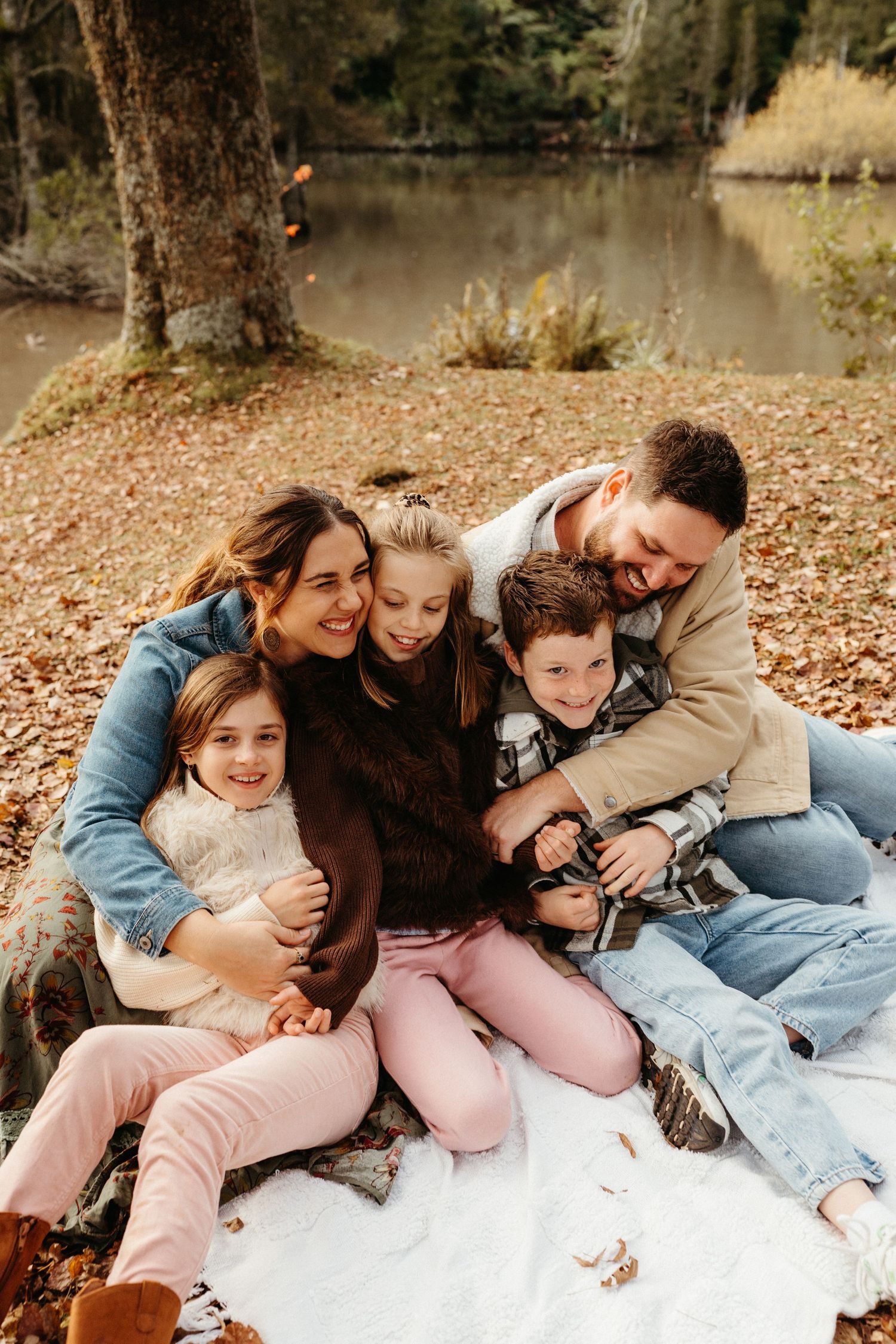 Happy family of five cuddling on a blanket by a serene lake surrounded by autumn foliage.