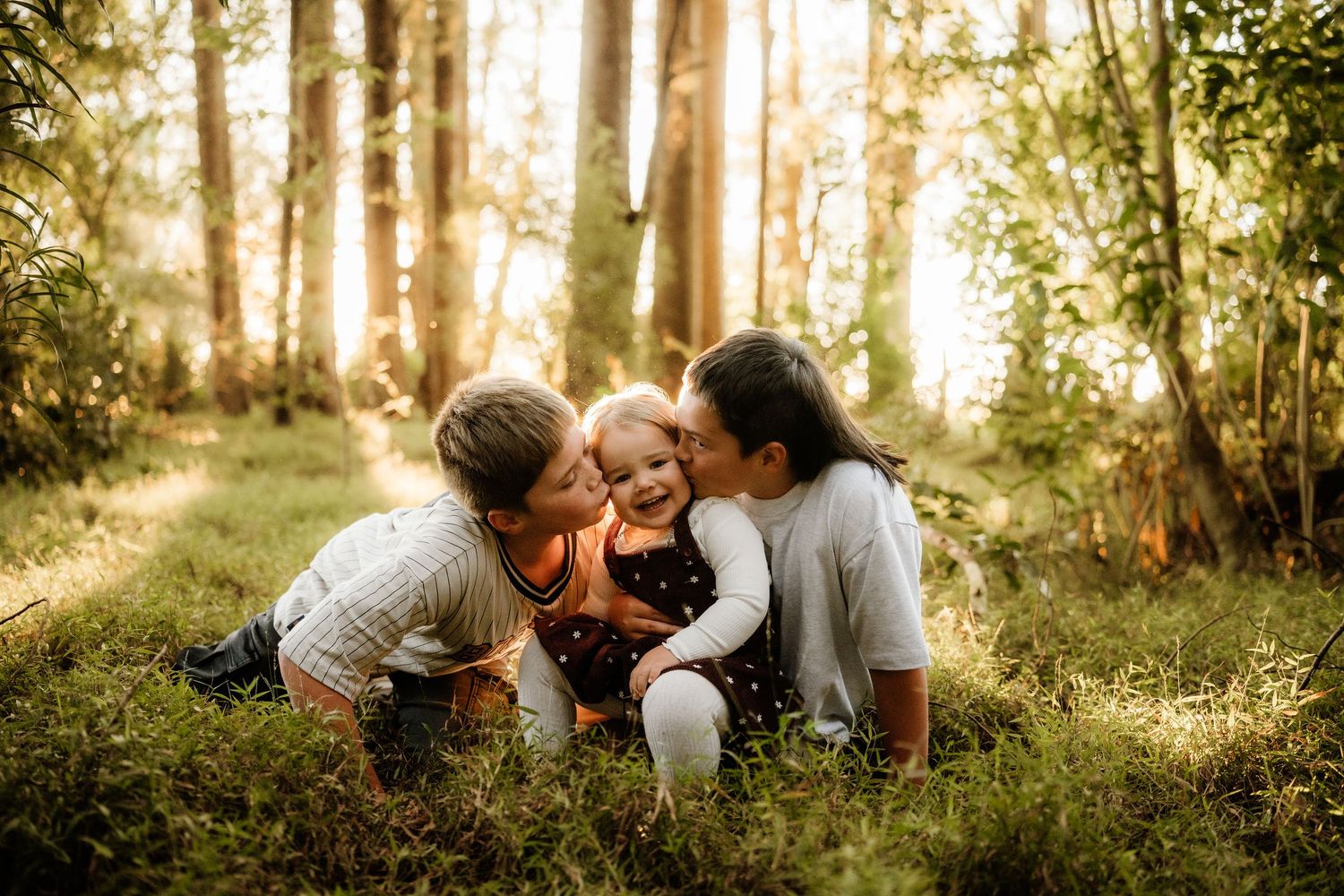 Two parents kissing their baby on the cheeks in a sunlit forest clearing with lush green grass.