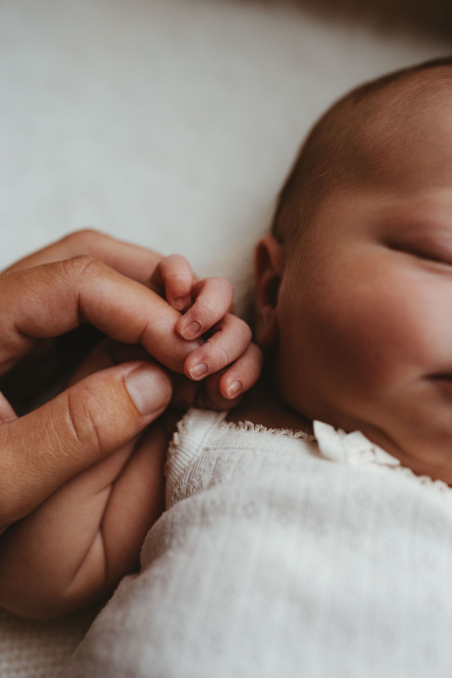 A sleeping newborn baby gently grips an adult finger, wrapped in a soft white blanket.