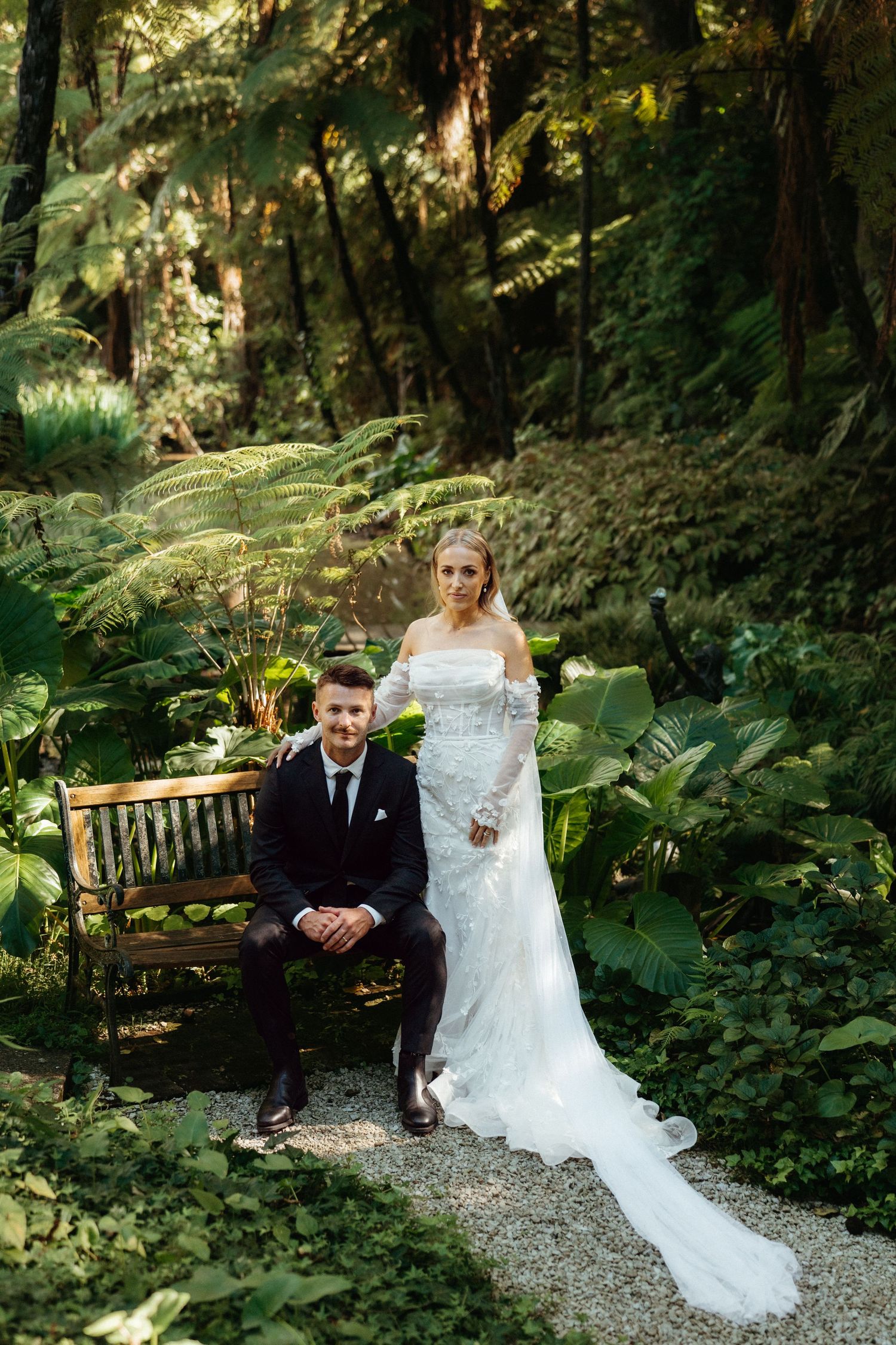 Bride in off-shoulder lace gown and groom in black suit posing in a lush green garden.