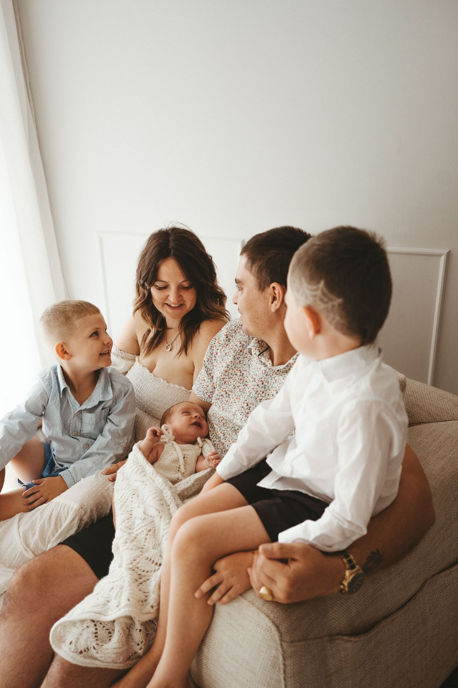 Family of five sitting on a cozy sofa, parents admiring their newborn baby while two young boys look on warmly.