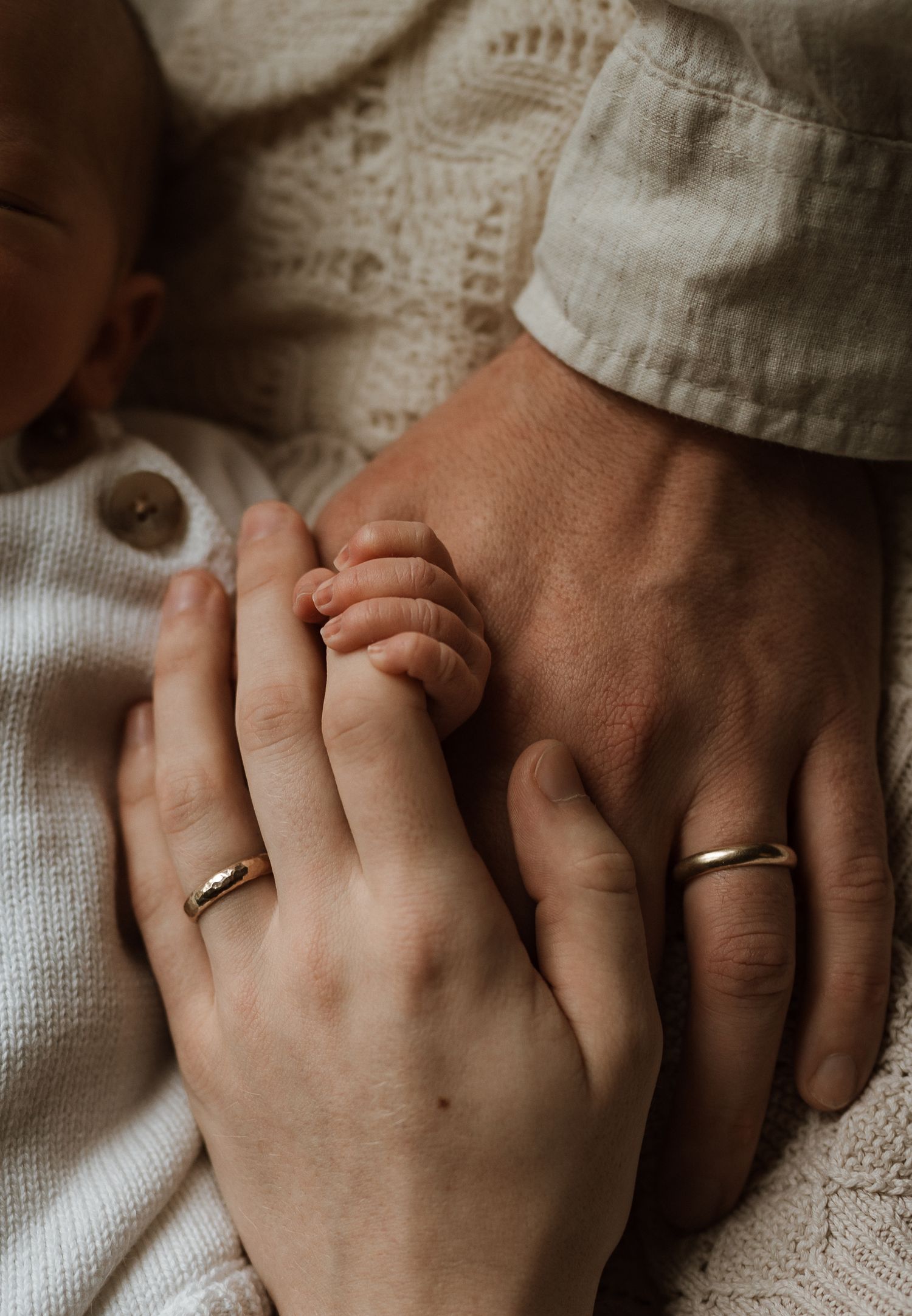 Two married parents with gold wedding bands gently hold their newborn baby's tiny hand in a tender close-up photo.