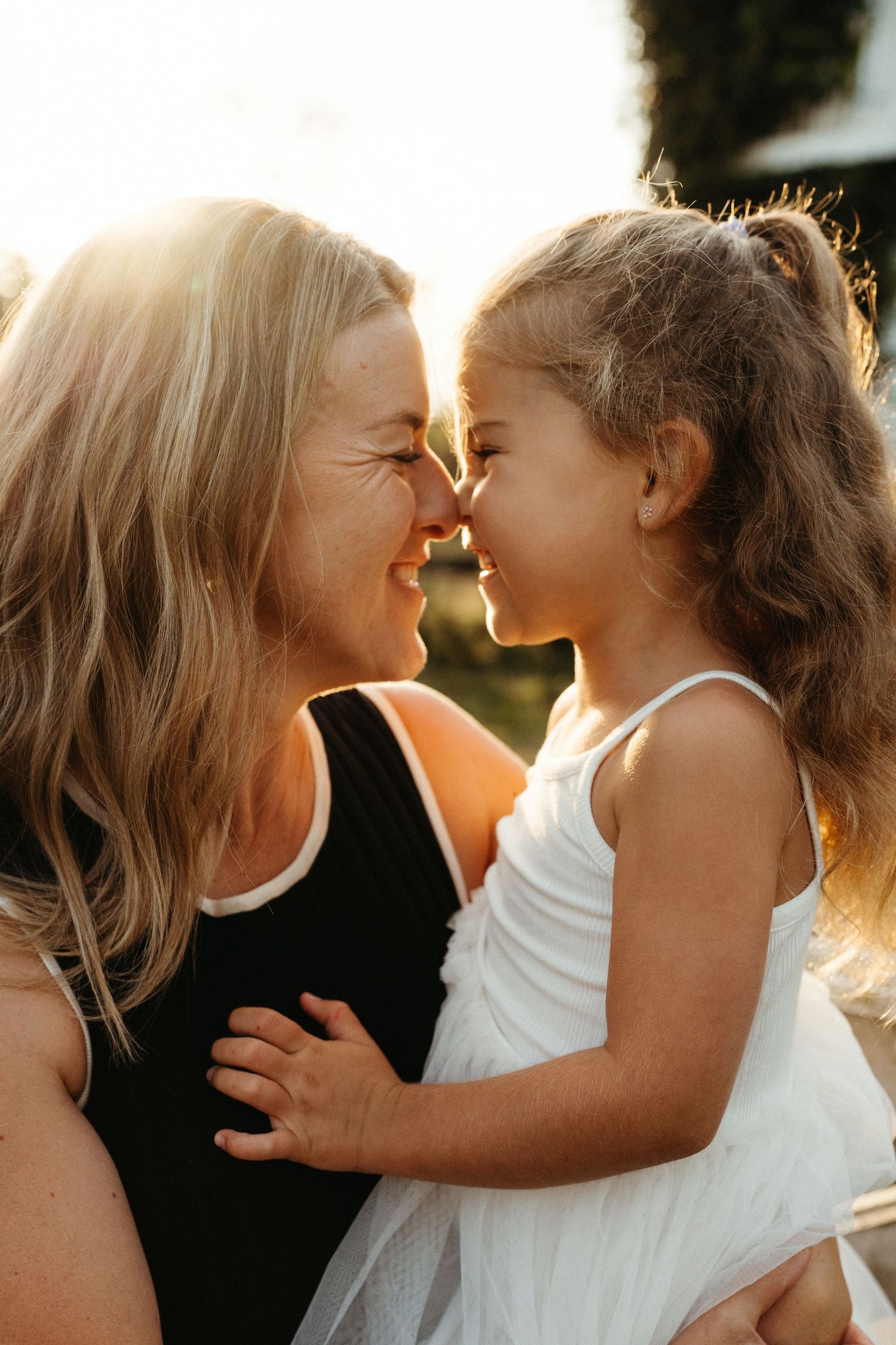 A smiling woman holds a young girl as they touch noses in a warm golden sunset light outdoors.