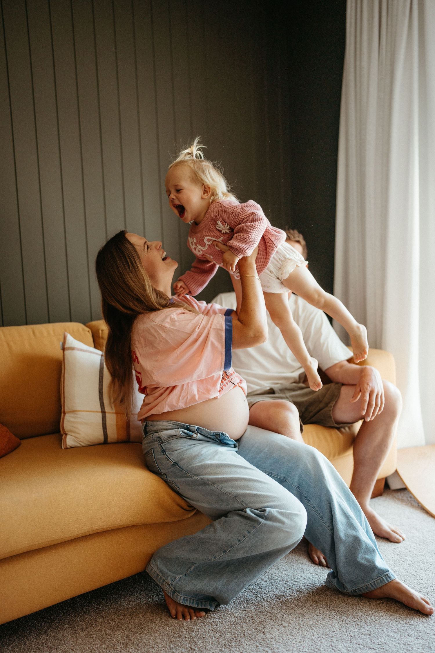 Pregnant woman lifting laughing toddler girl on yellow sofa at home, joyful family moment indoors.