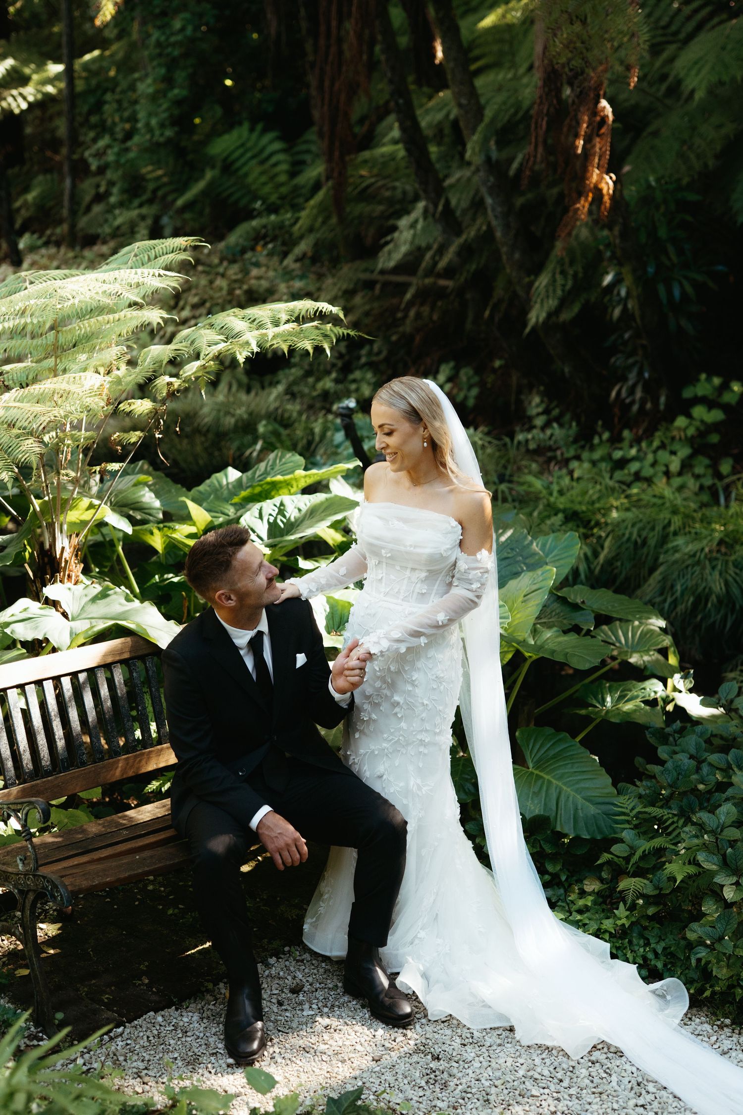 Bride in fitted lace gown and groom in black suit sharing a tender moment on a garden bench.
