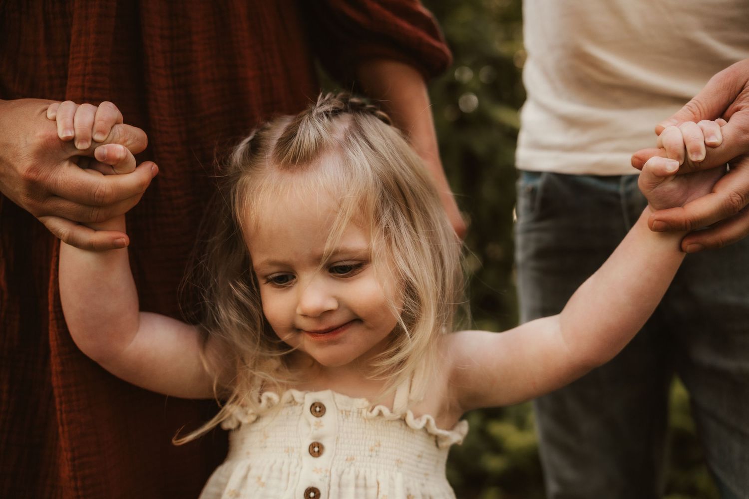 Toddler girl in white dress holds hands with parents outdoors, smiling joyfully in a warm family moment. Katie Charlotte Photography