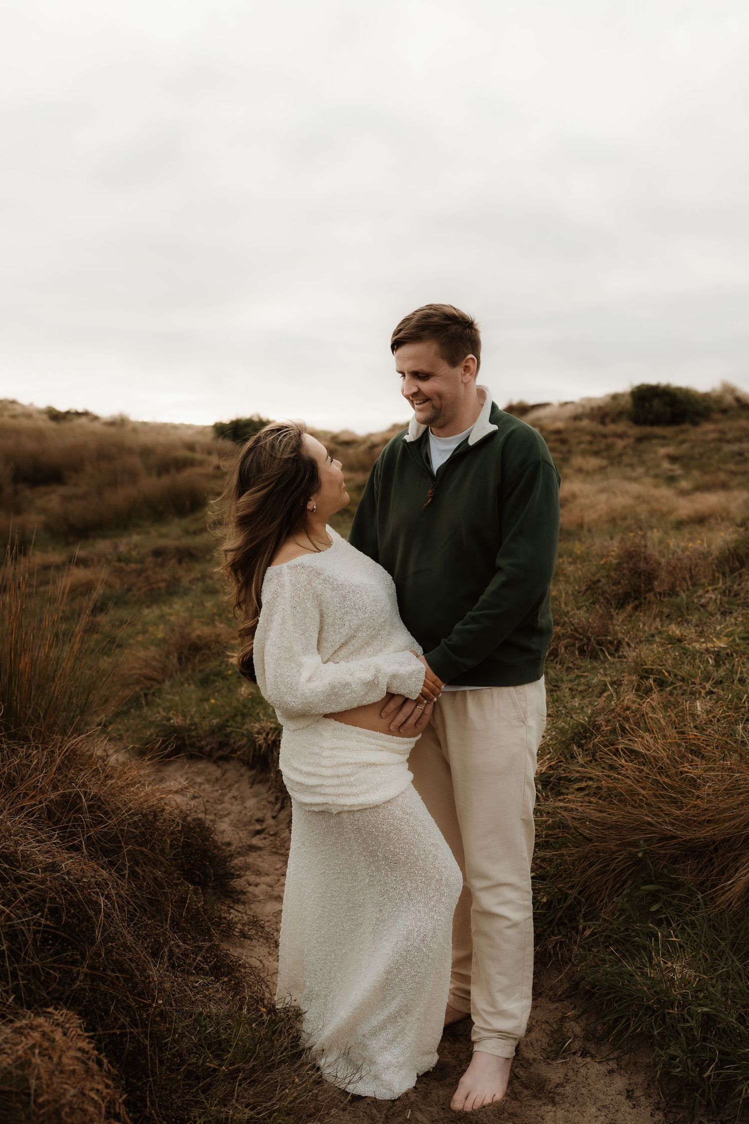Couple sharing a tender moment outdoors, pregnant woman in white dress with partner in green sweater among golden grasses.