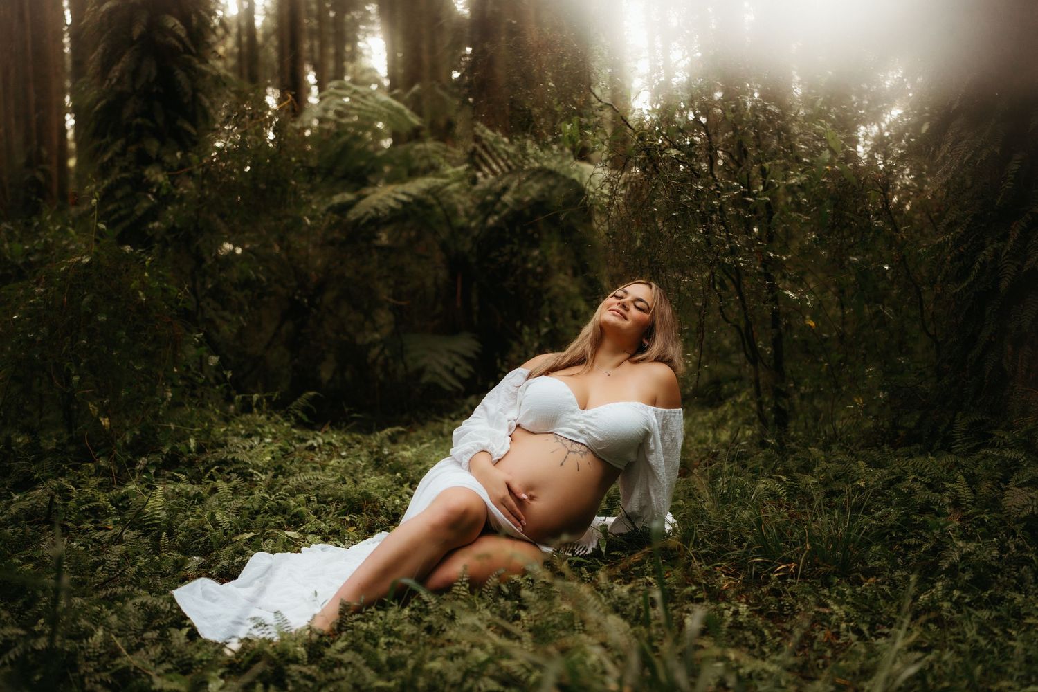 Pregnant woman in white lingerie sitting peacefully in a lush green forest, cradling her baby bump in soft natural light.