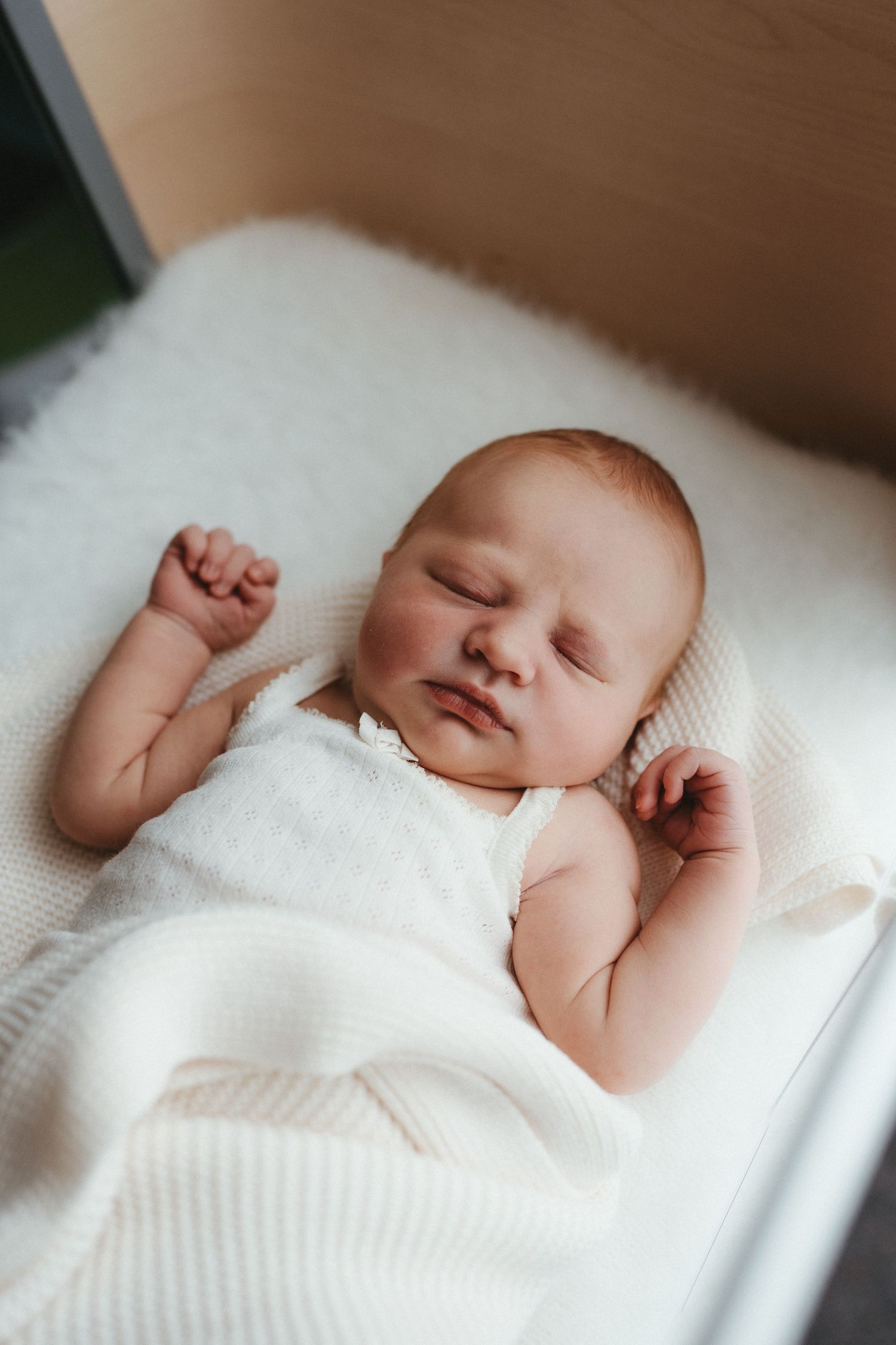 Newborn baby sleeping peacefully in white bassinet with arms raised in white wrap.