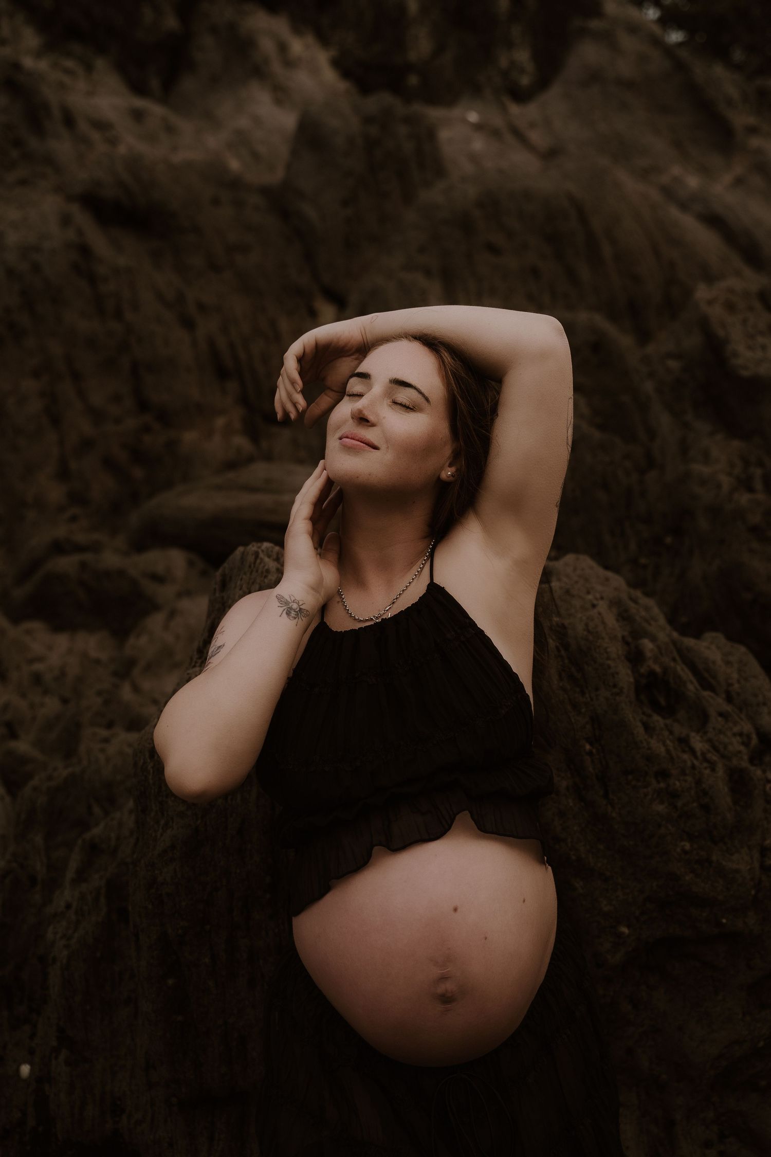 Pregnant woman in black crop top poses gracefully against dark rocks, eyes closed, arm raised, showcasing baby bump outdoors.