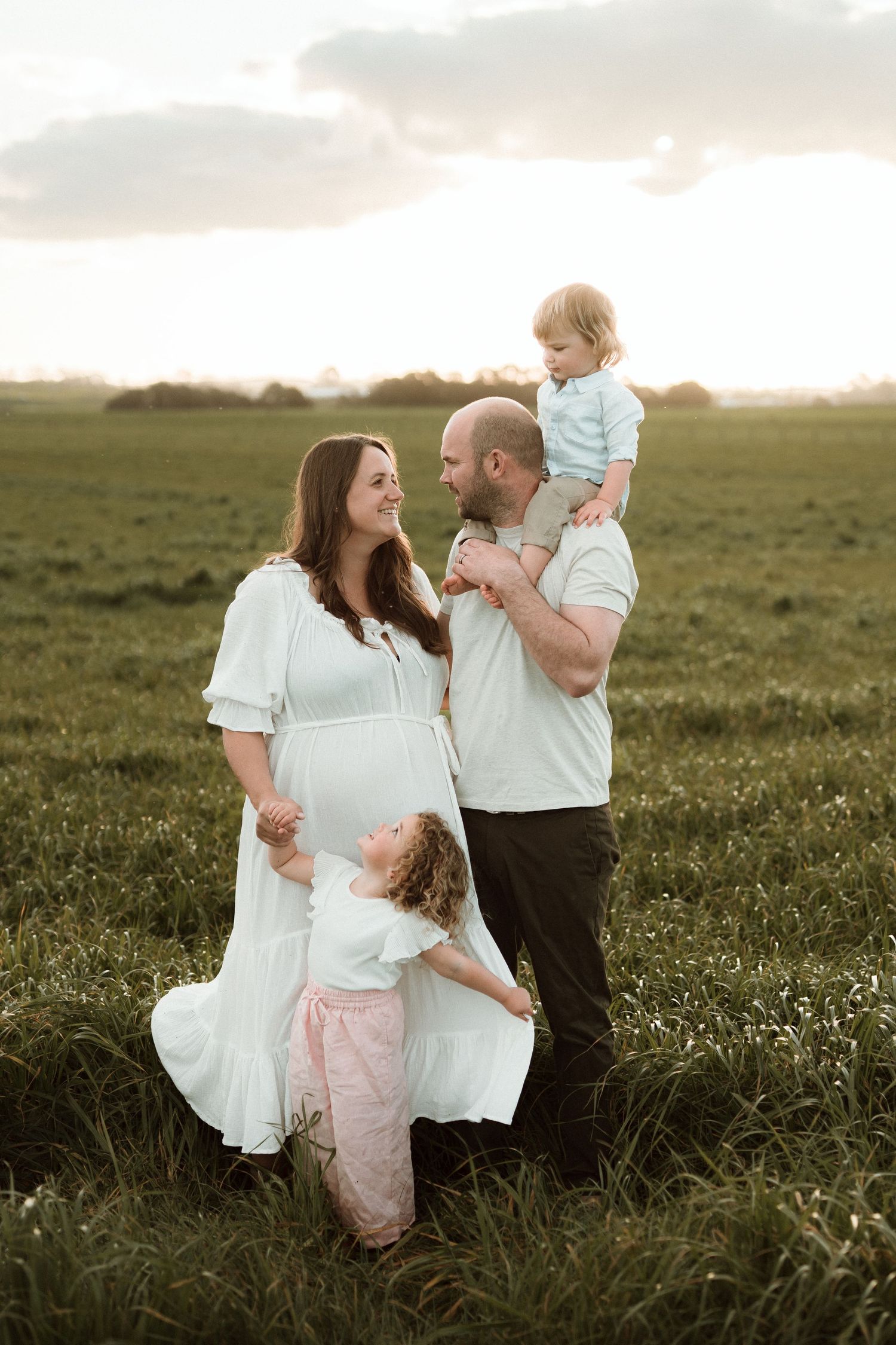 Pregnant woman in white dress with husband and two young children in a green field at golden hour. Katie Charlotte Photography