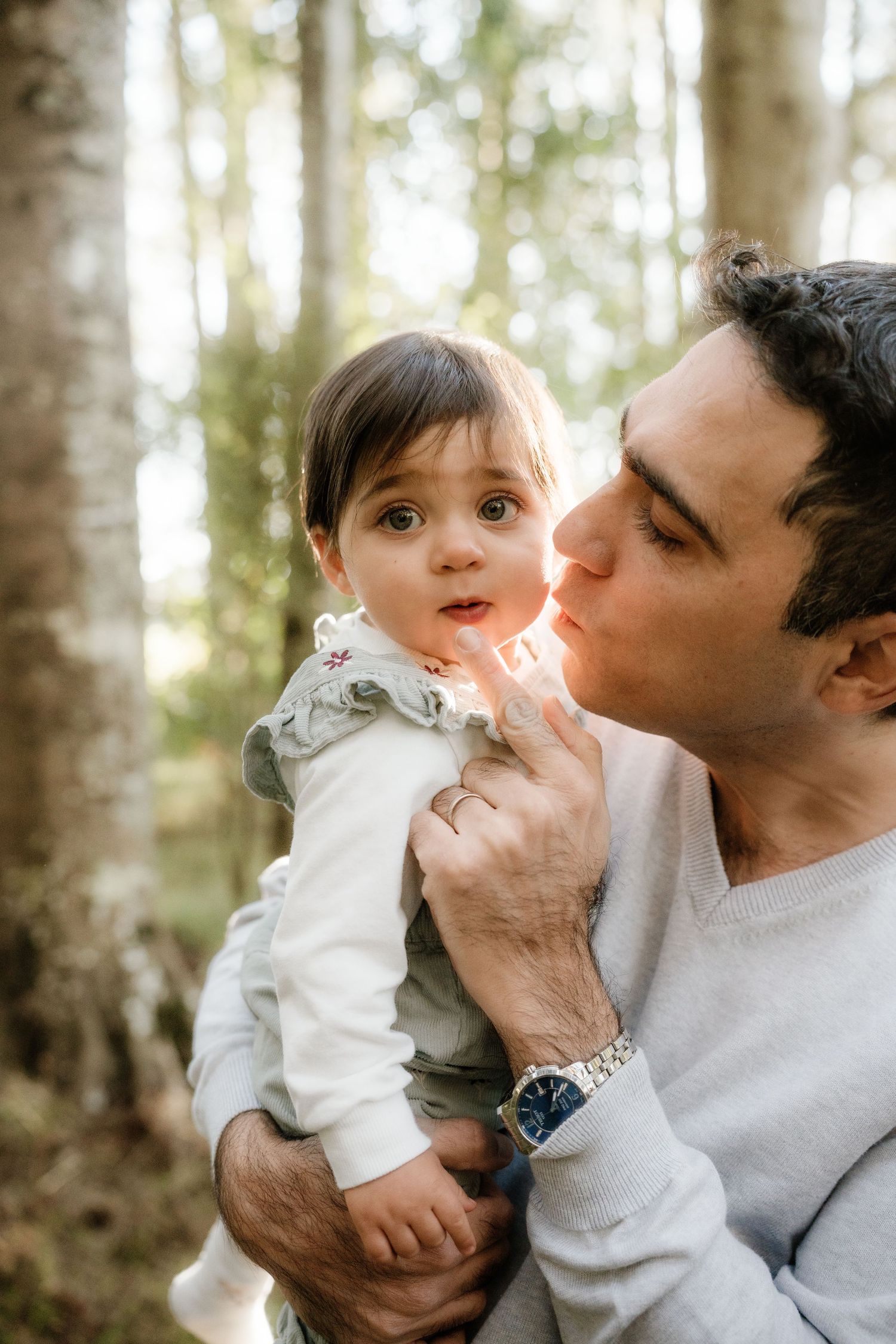 A father tenderly kisses his baby girl's cheek while holding her in a sunlit forest setting.