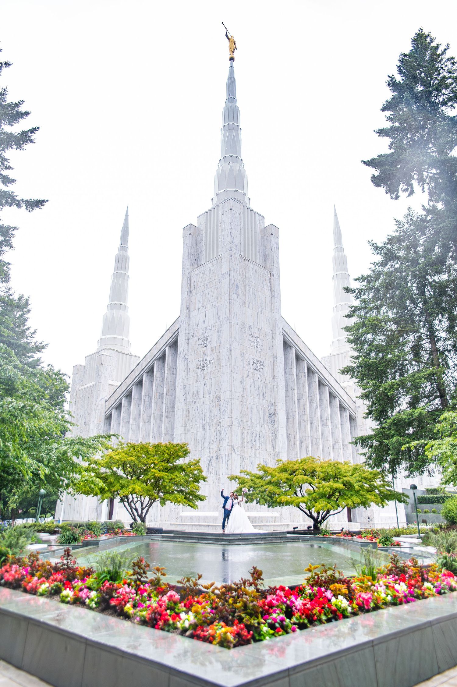 Mormon temple with white spires surrounded by vibrant flower gardens and trees.