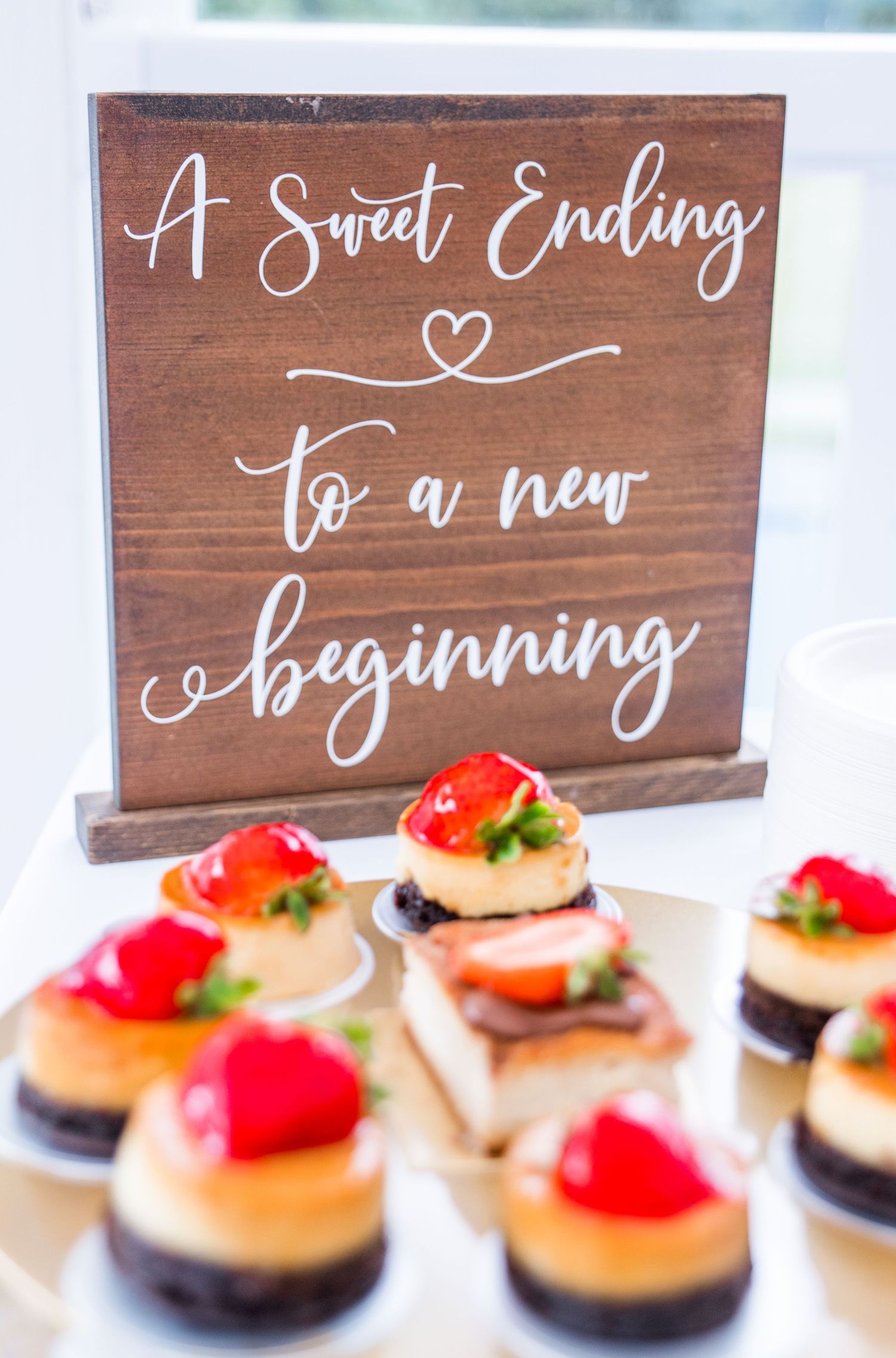 Wedding dessert table with mini cheesecakes topped with strawberries beside a wooden sign reading A Sweet Ending to a new beginning.
