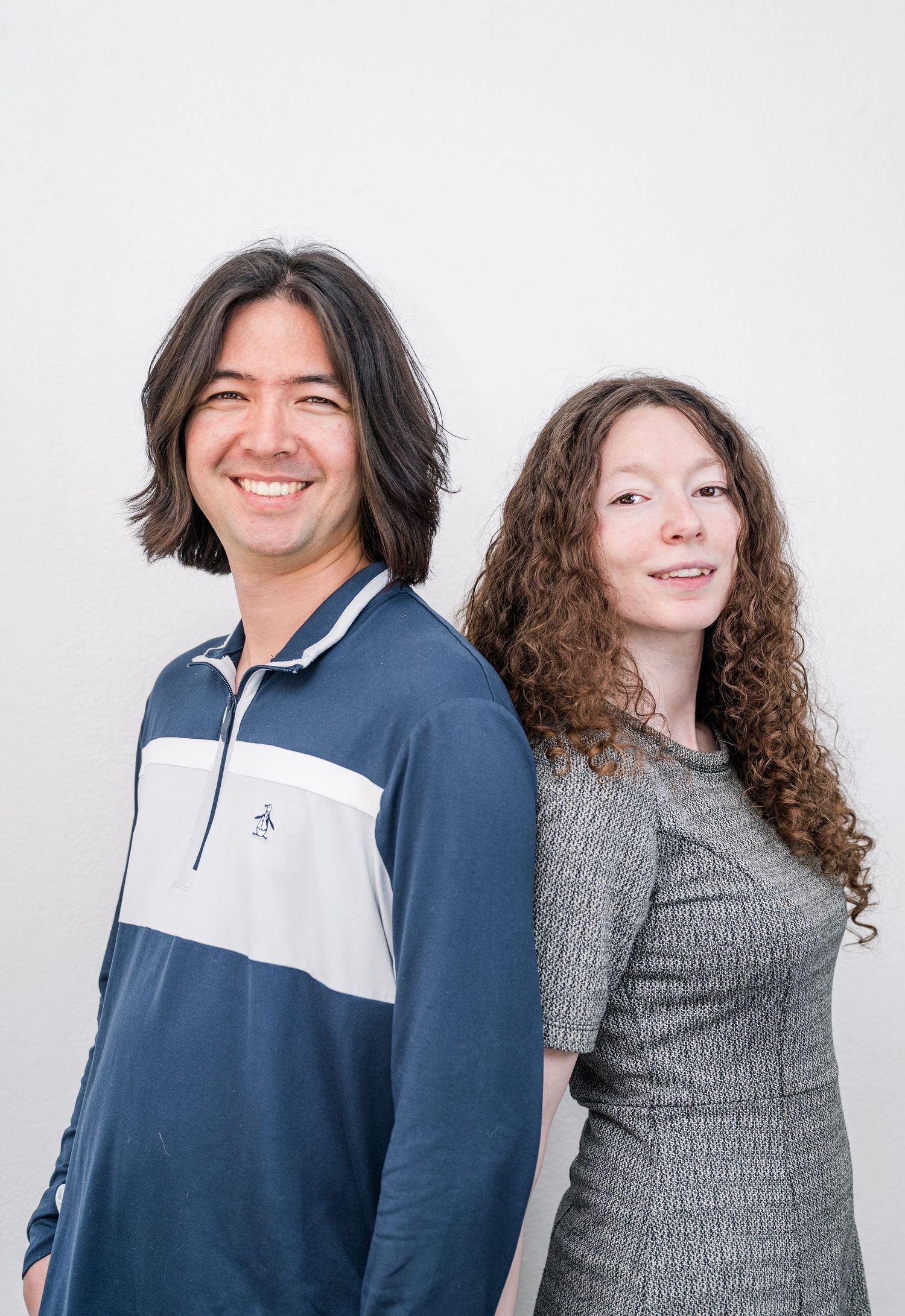 Two smiling people posing back-to-back, man in blue polo and woman in gray dress, against white background.