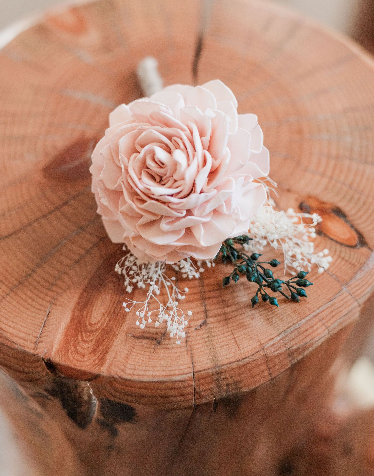 A delicate blush pink rose boutonniere with baby's breath and teal berries resting on a rustic wood stump.