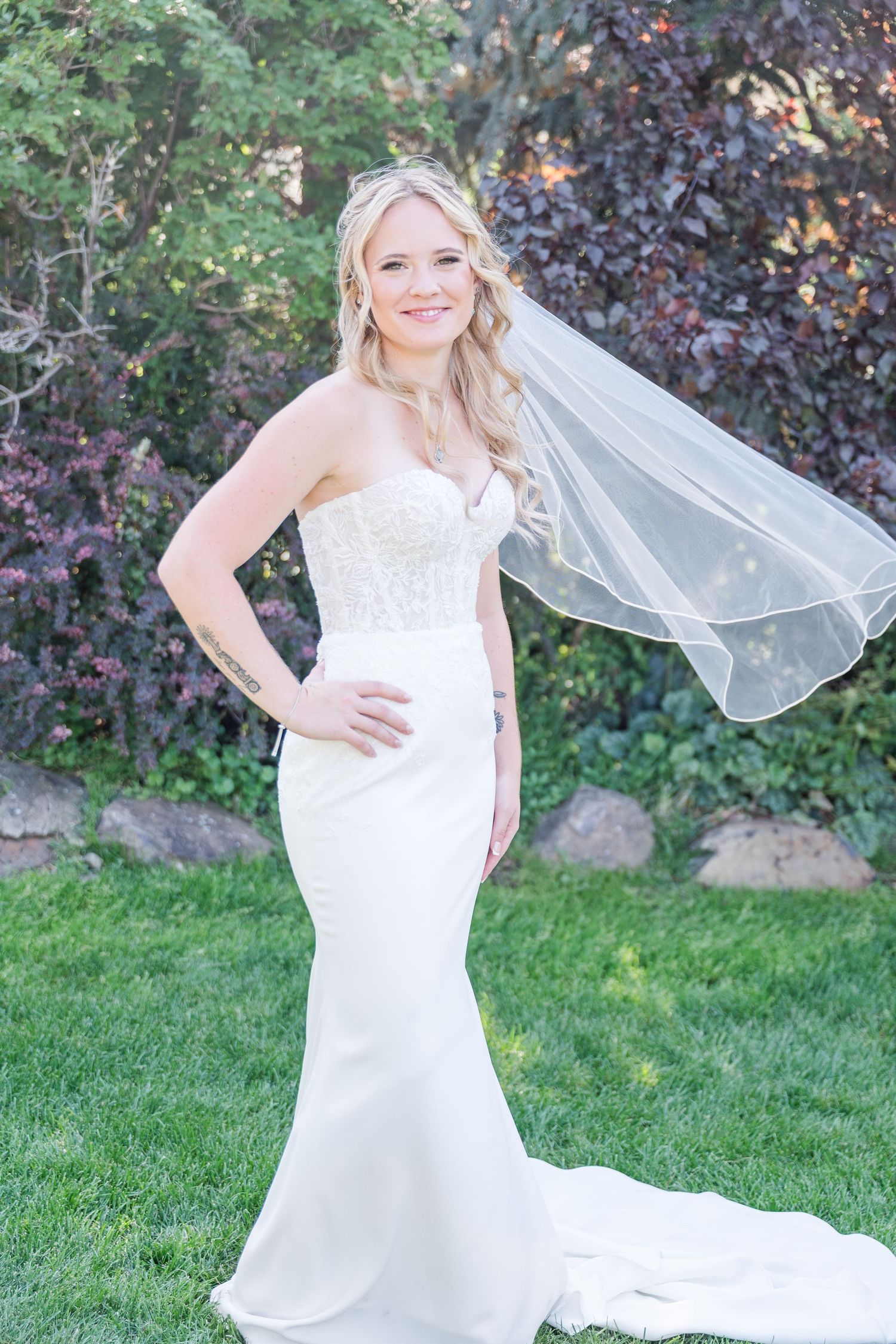 Smiling bride in strapless lace mermaid gown with flowing veil standing on green lawn surrounded by lush garden.