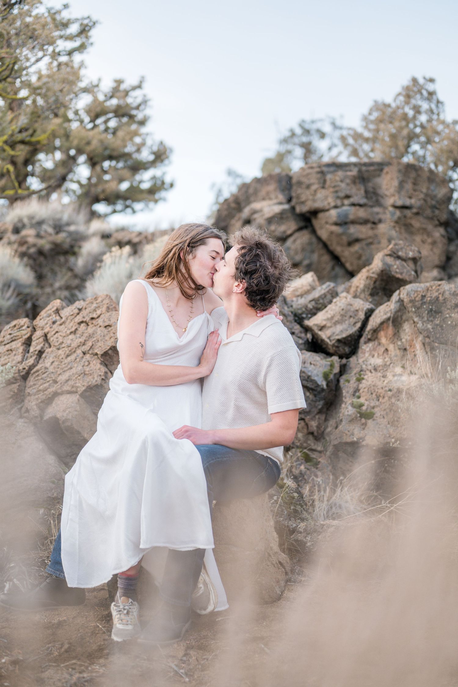 Couple sharing a romantic kiss on rocky terrain, woman in white dress, man in cream shirt, with desert shrubs and boulders.