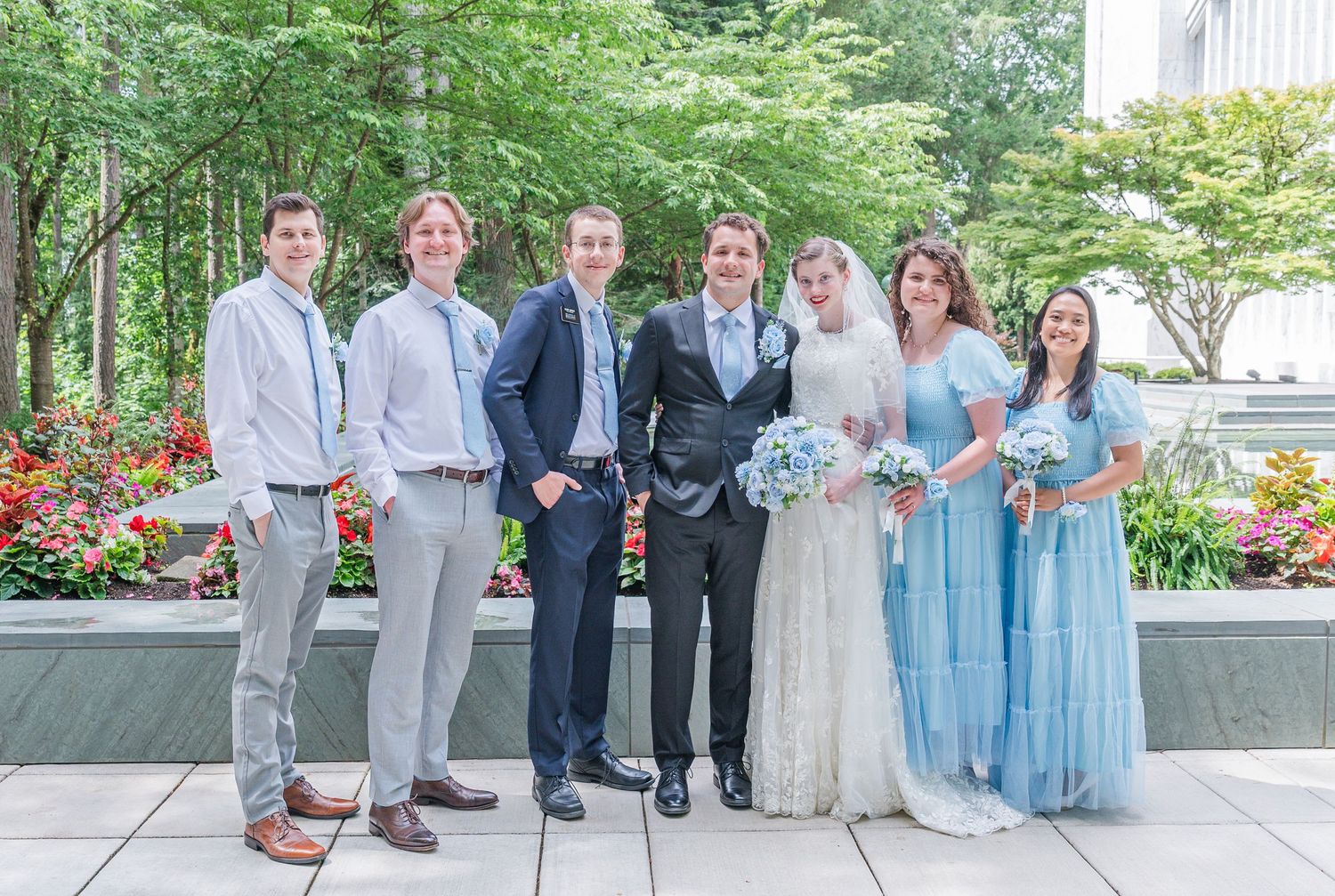 Wedding party posing outdoors, bride and groom with bridesmaids in blue dresses and groomsmen in suits, surrounded by greenery.