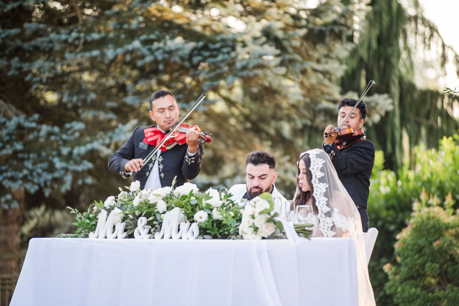 Two mariachi violinists serenade a couple at a floral wedding reception table outdoors.