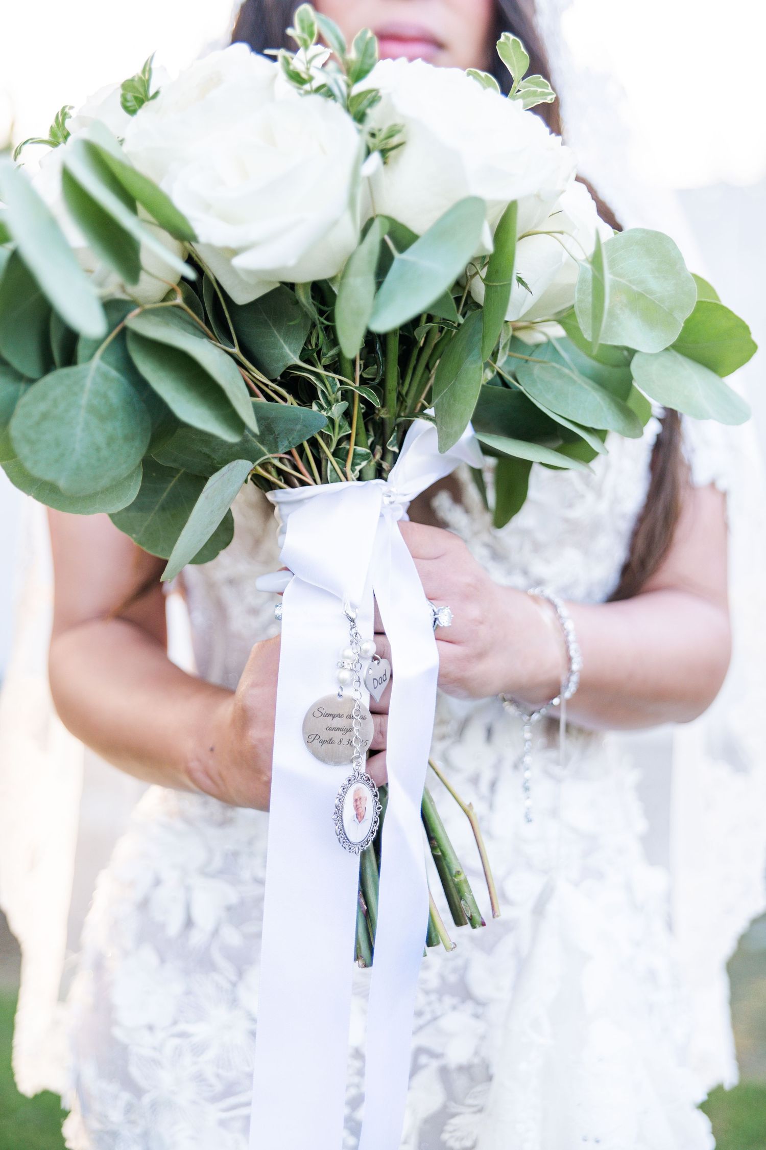Bride holding white rose bouquet with green eucalyptus leaves, tied with white ribbon and charm accents, in lace wedding dress.