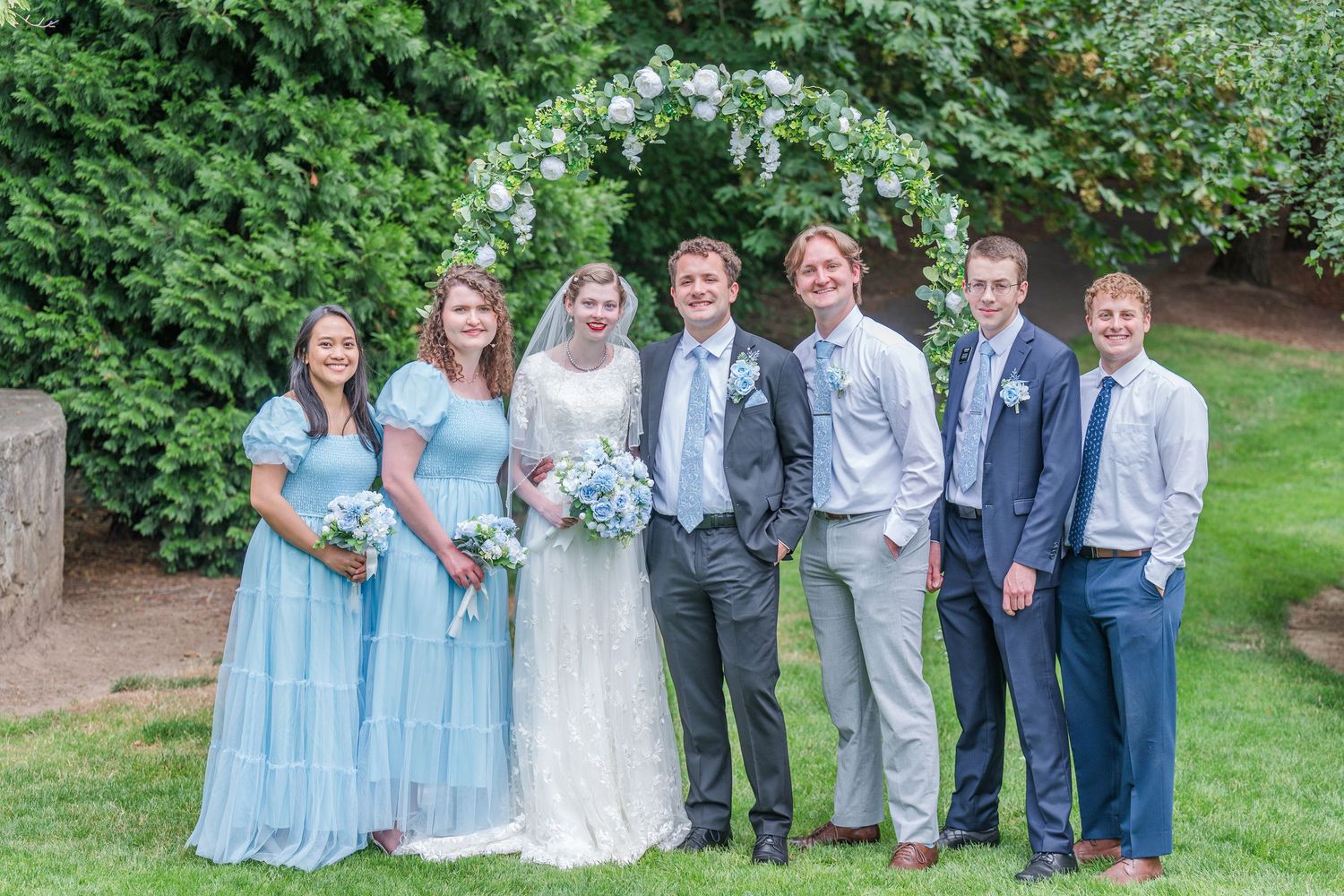 Wedding party poses under floral arch, bride in white gown with bridesmaids in blue dresses and groomsmen in suits.