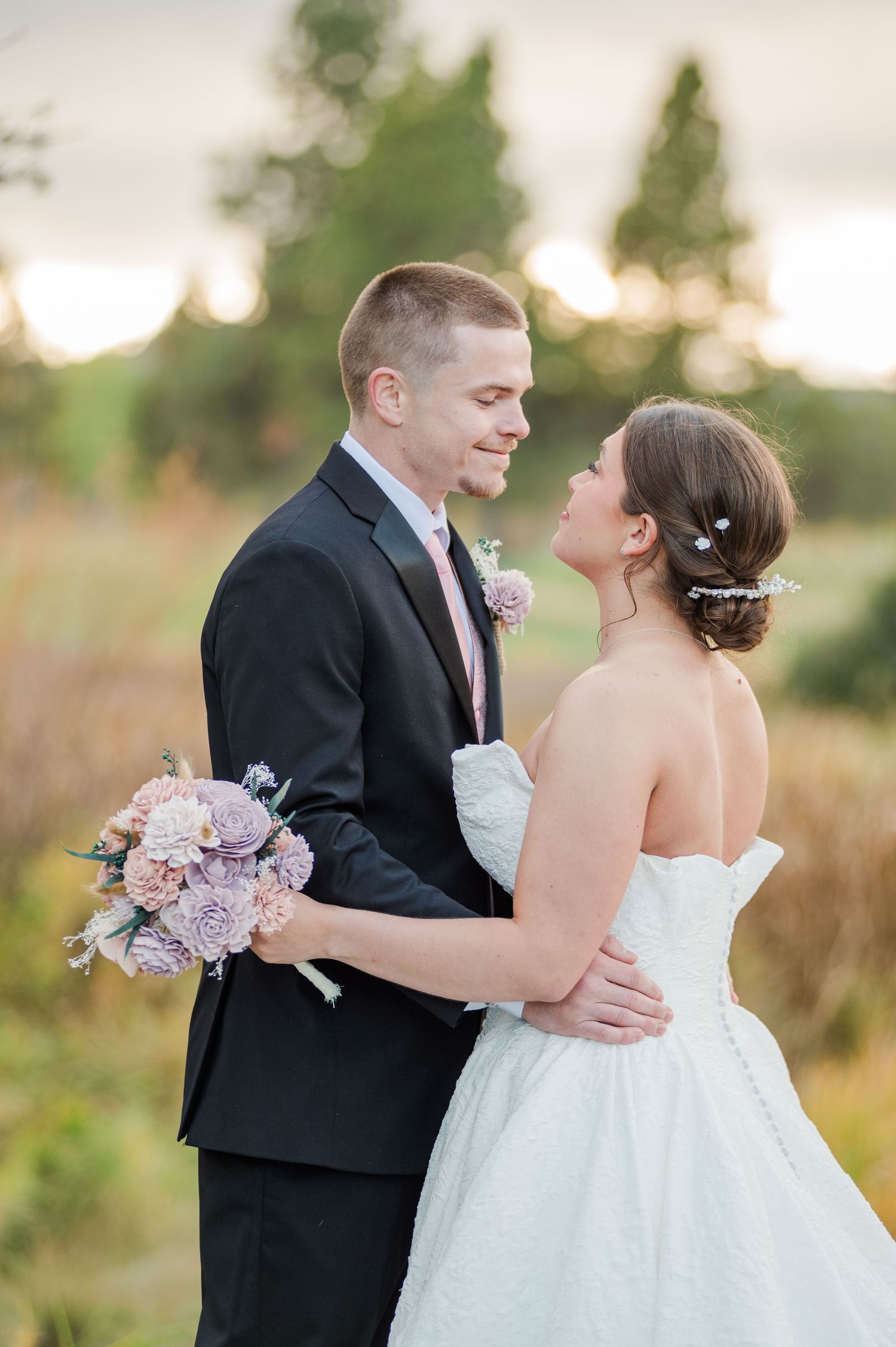 Bride and groom share a romantic moment outdoors at golden hour, she holds pink bouquet wearing strapless gown, he wears dark suit.