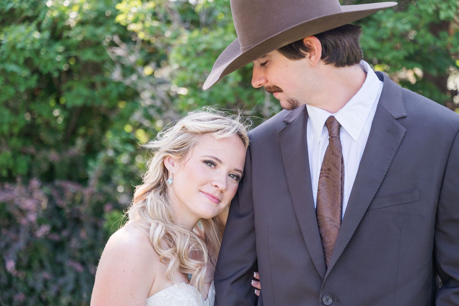 Bride in white dress leans on groom wearing gray suit and brown cowboy hat outdoors at wedding.