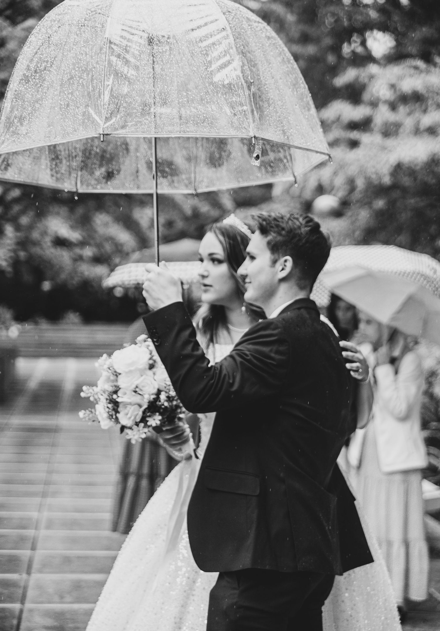 Wedding party holding umbrellas in the rain during outdoor ceremony.