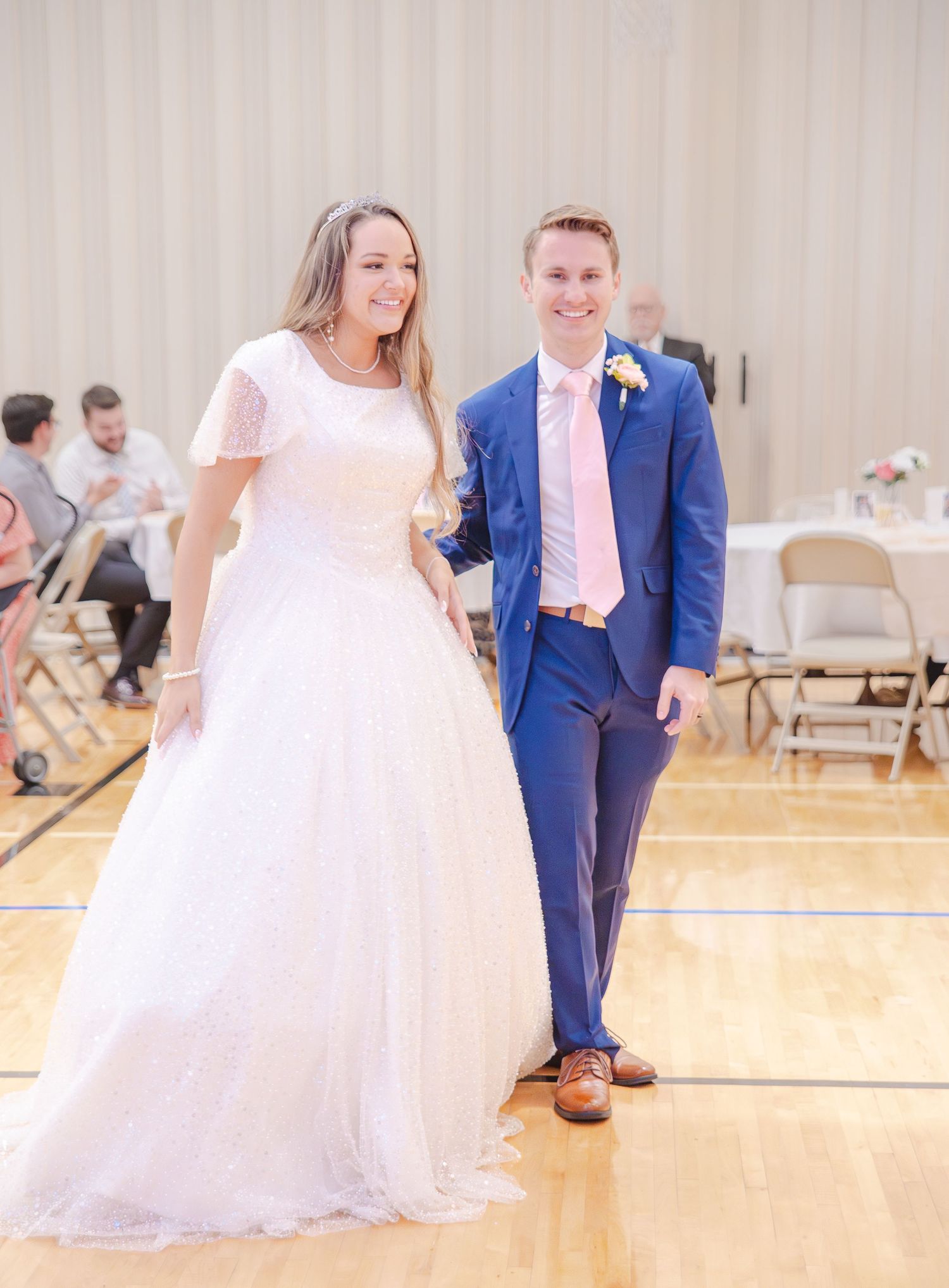 Wedding couple dances together in a bright reception hall with wooden floors and folding chairs.