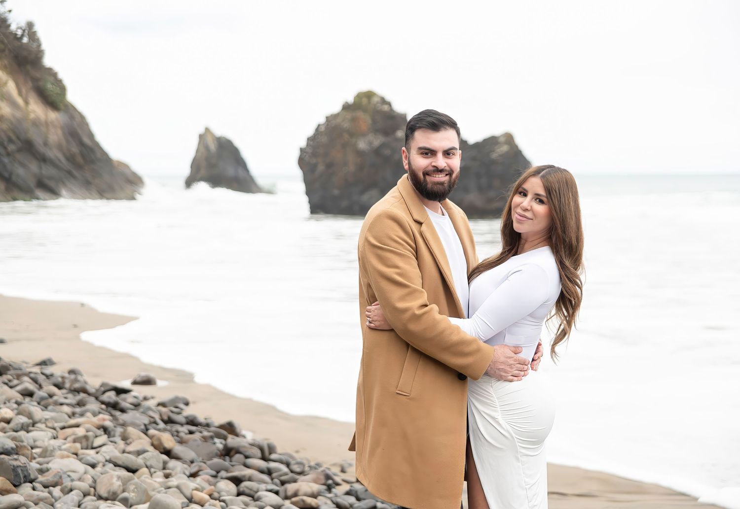 Couple shares romantic moments on rocky beach coastline in casual formal wear.