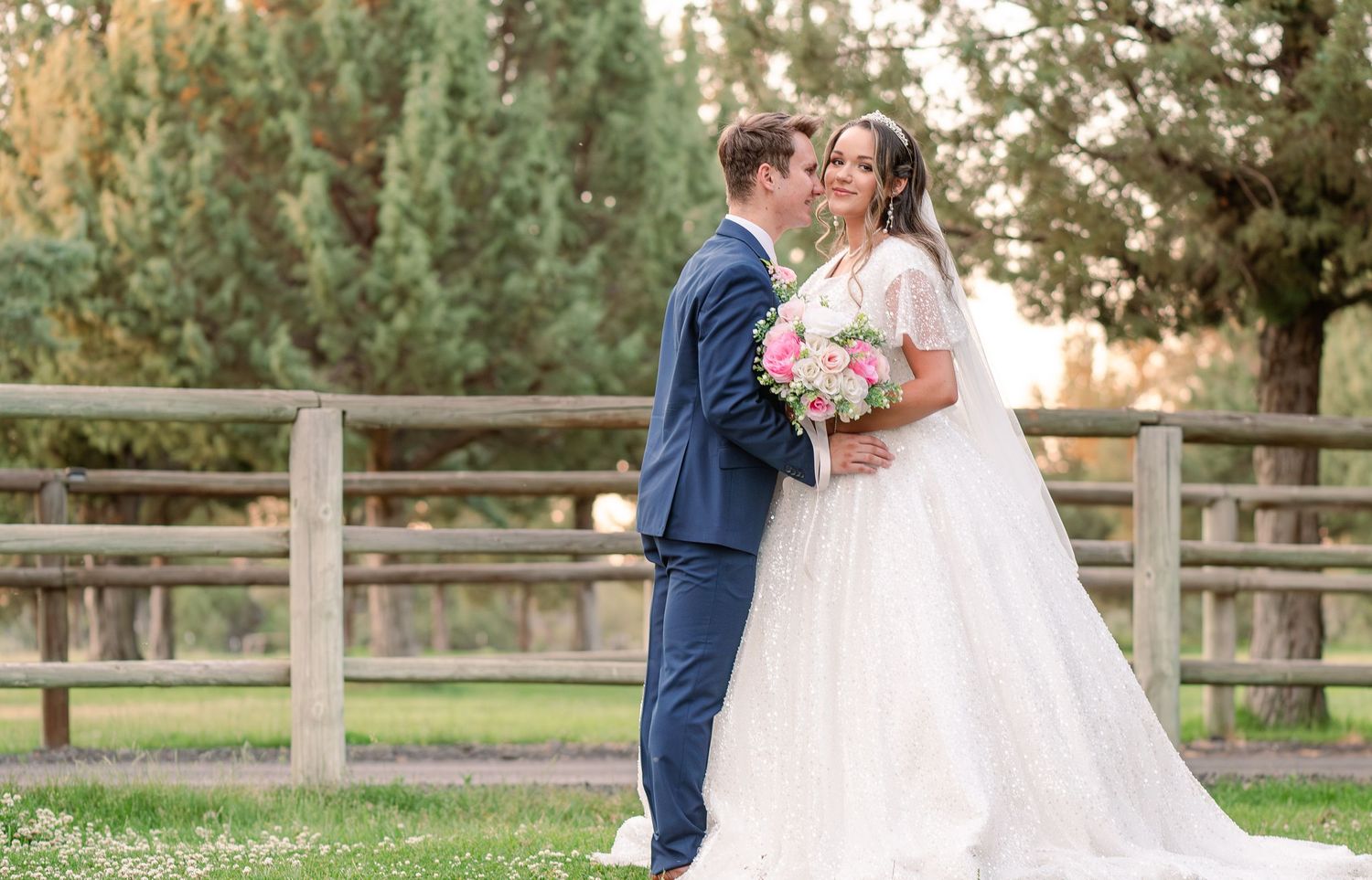 Wedding couple poses along rustic wooden fence in garden setting during golden hour.