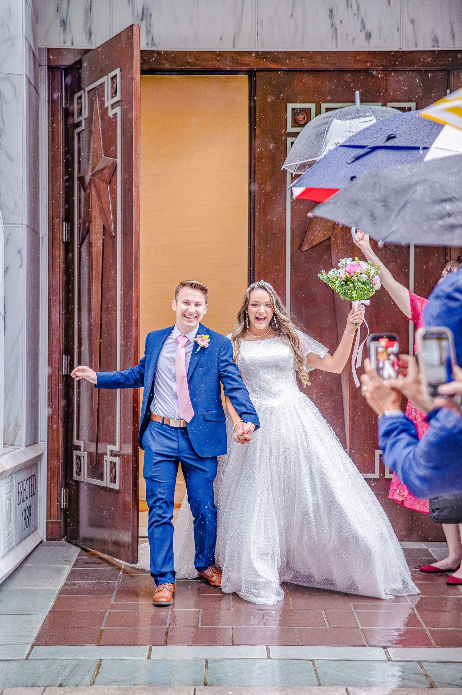Wedding party exiting a brick building with umbrellas on a rainy day.