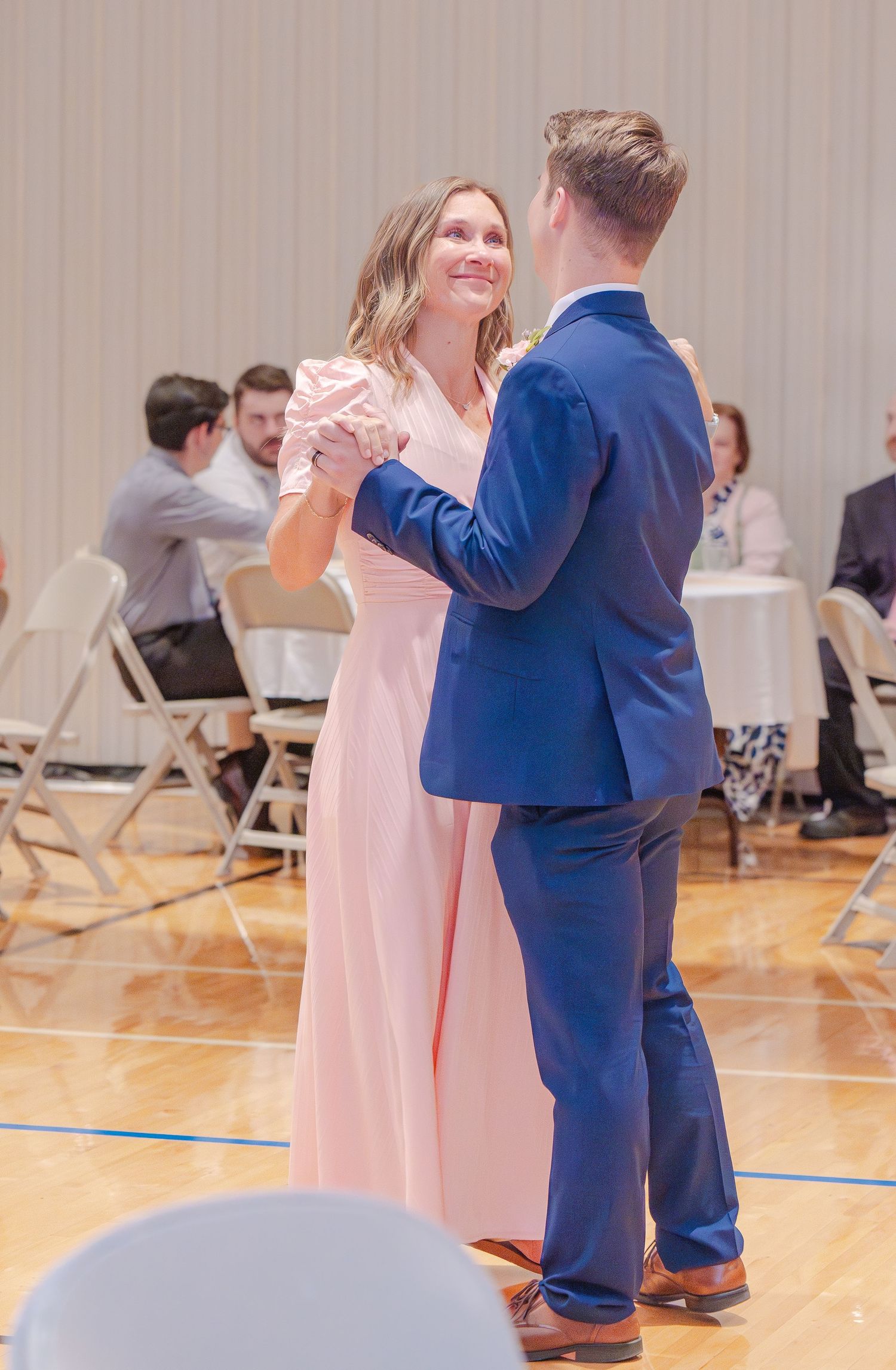 Wedding party members in blue and pink attire dance together on a gymnasium floor.