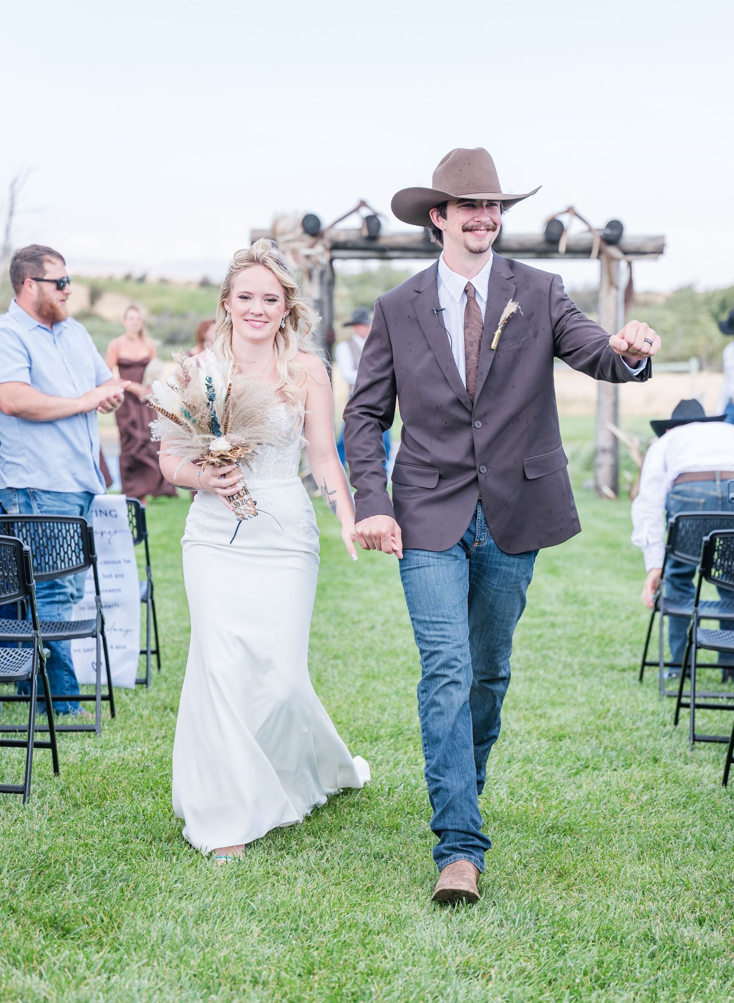 Newlywed couple walks down outdoor wedding aisle, groom in cowboy hat and brown suit, bride in white gown holding bouquet.