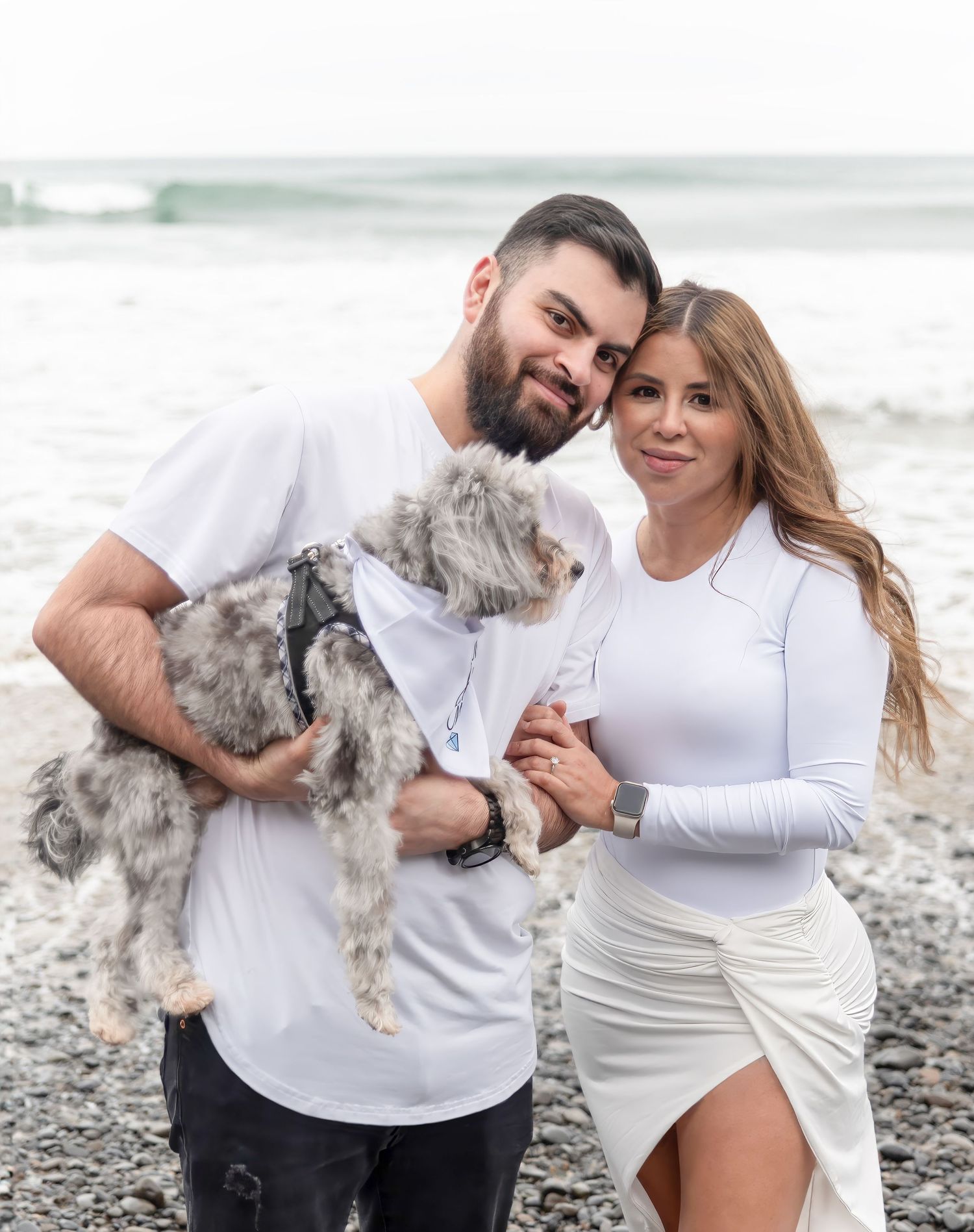 Couple in matching white outfits holding a fluffy gray dog on a rocky beach with ocean waves in the background.