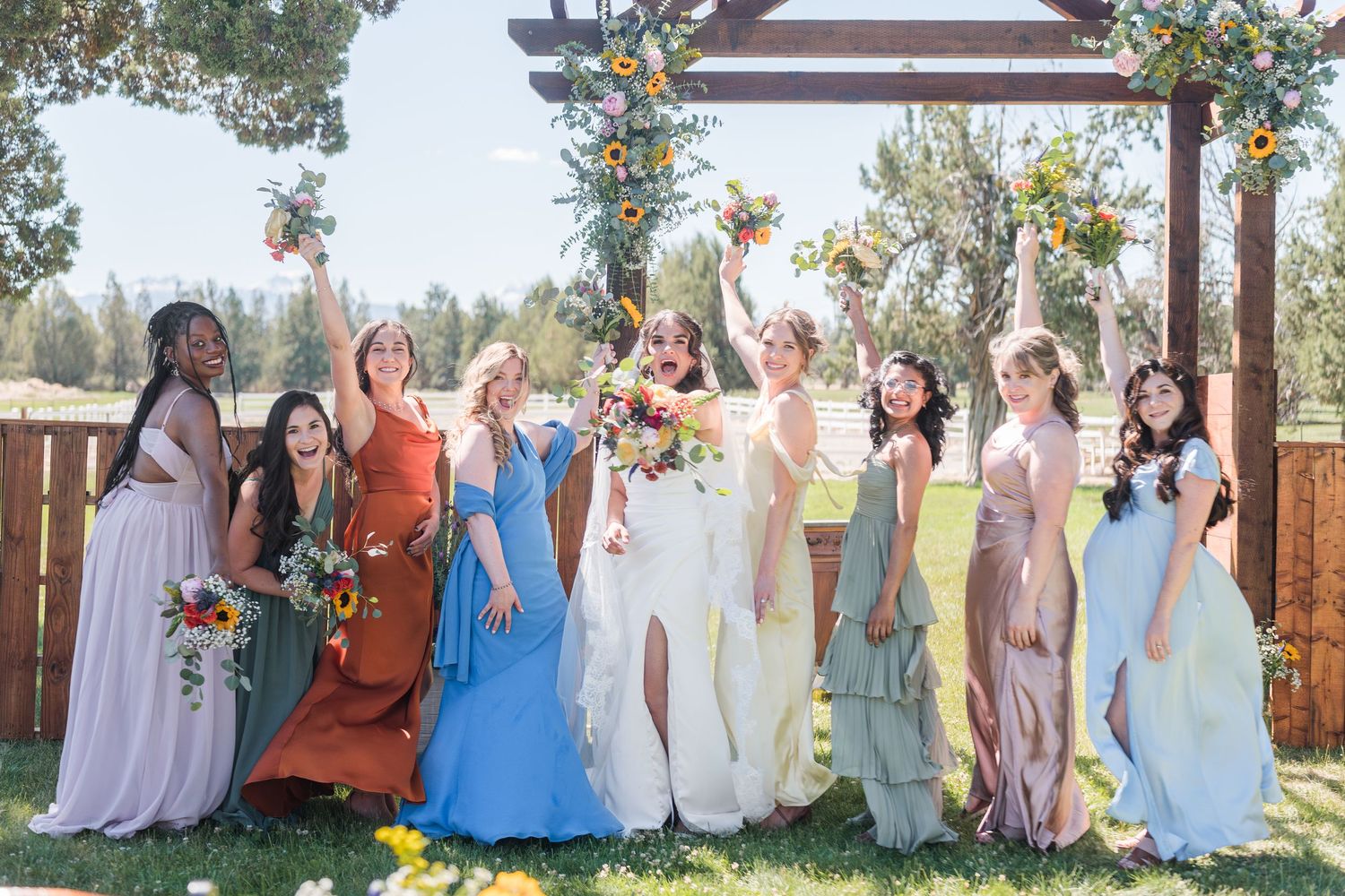 Bride in white gown poses with colorful bridesmaids raising bouquets outdoors under floral arch on sunny day.